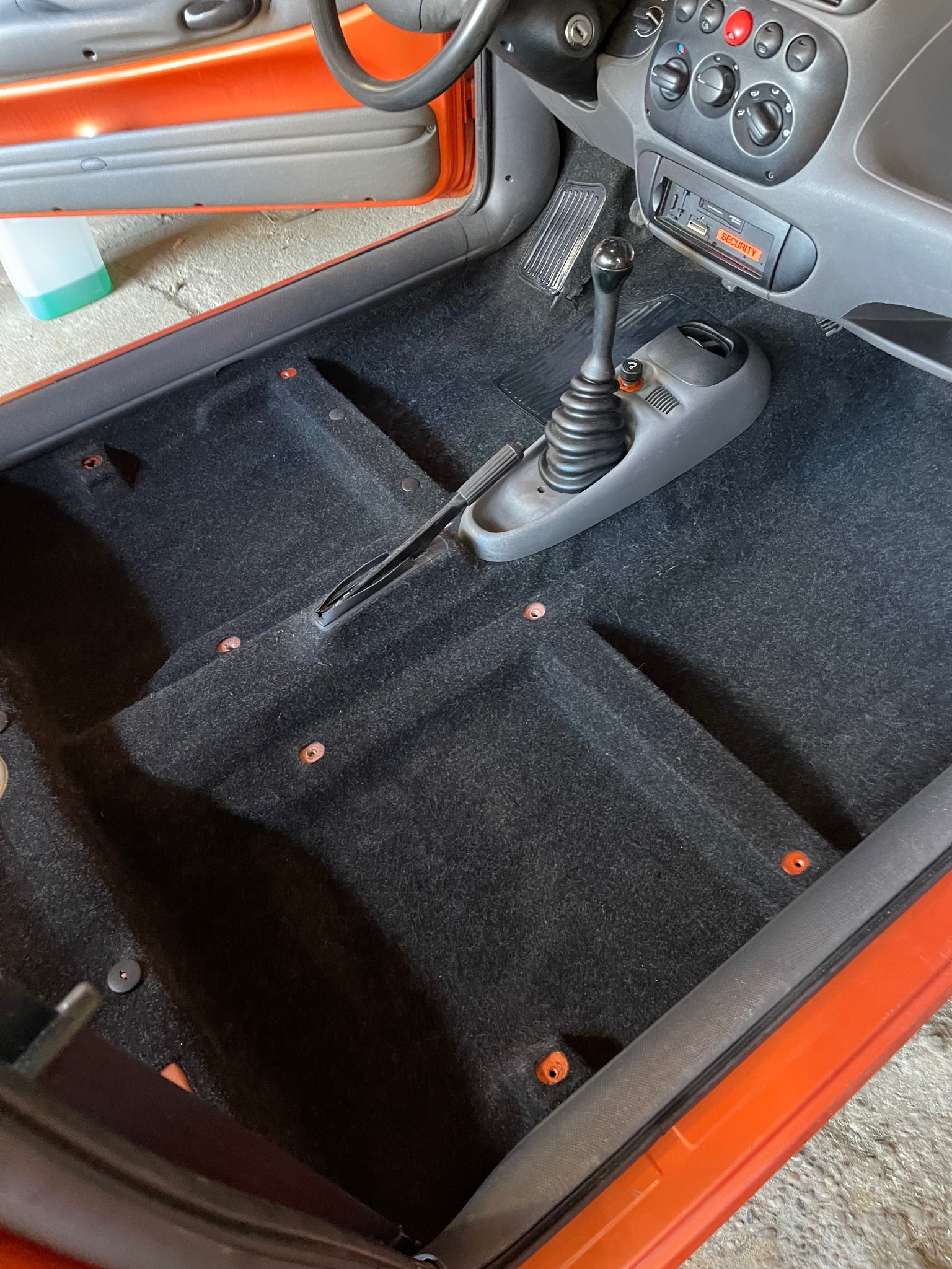 Wnętrze pomarańczowego samochodu z czarną wykładziną podłogową, widoczna dźwignia zmiany biegów, kokpit i uchylone drzwi, przygotowane do czyszczenia.