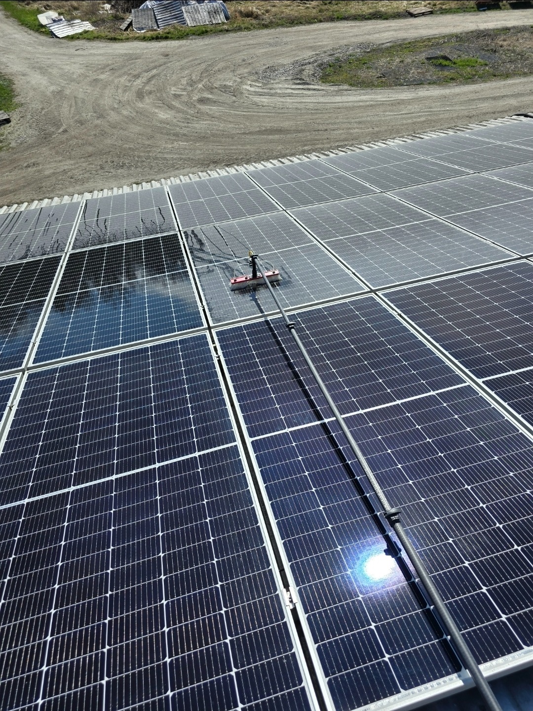 Czyszczenie paneli fotowoltaicznych szczotką teleskopową. Część paneli umyta, widać różnicę. W tle polna droga i budynki gospodarcze.