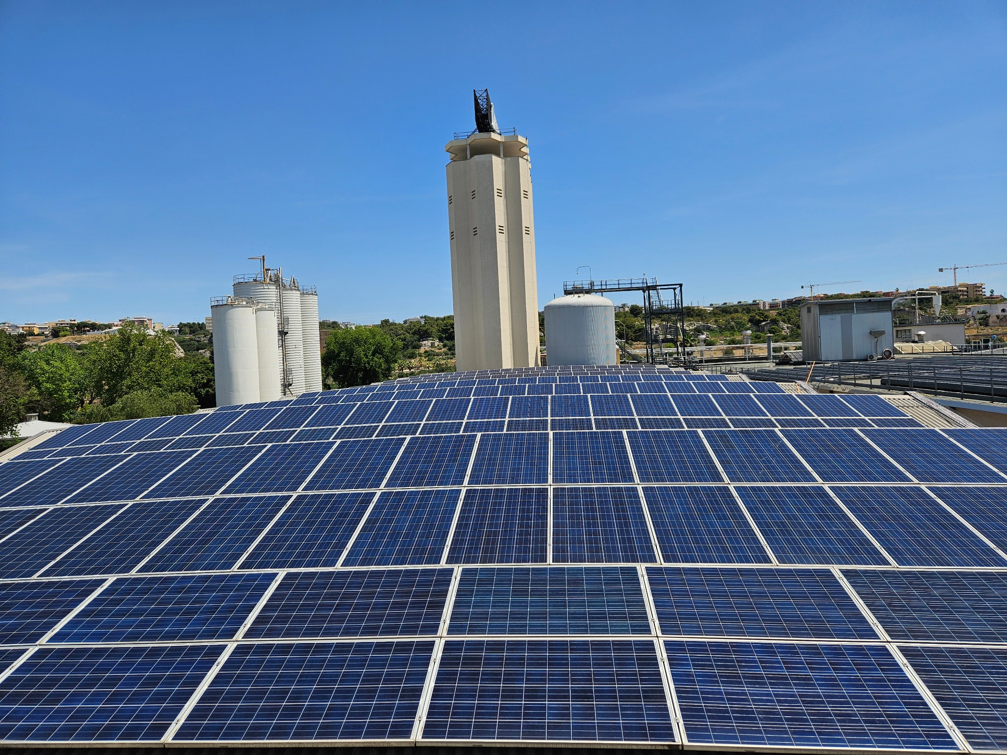 Rozległa instalacja paneli słonecznych na dachu budynku przemysłowego z widocznymi zbiornikami i wieżą w tle, na tle błękitnego nieba.