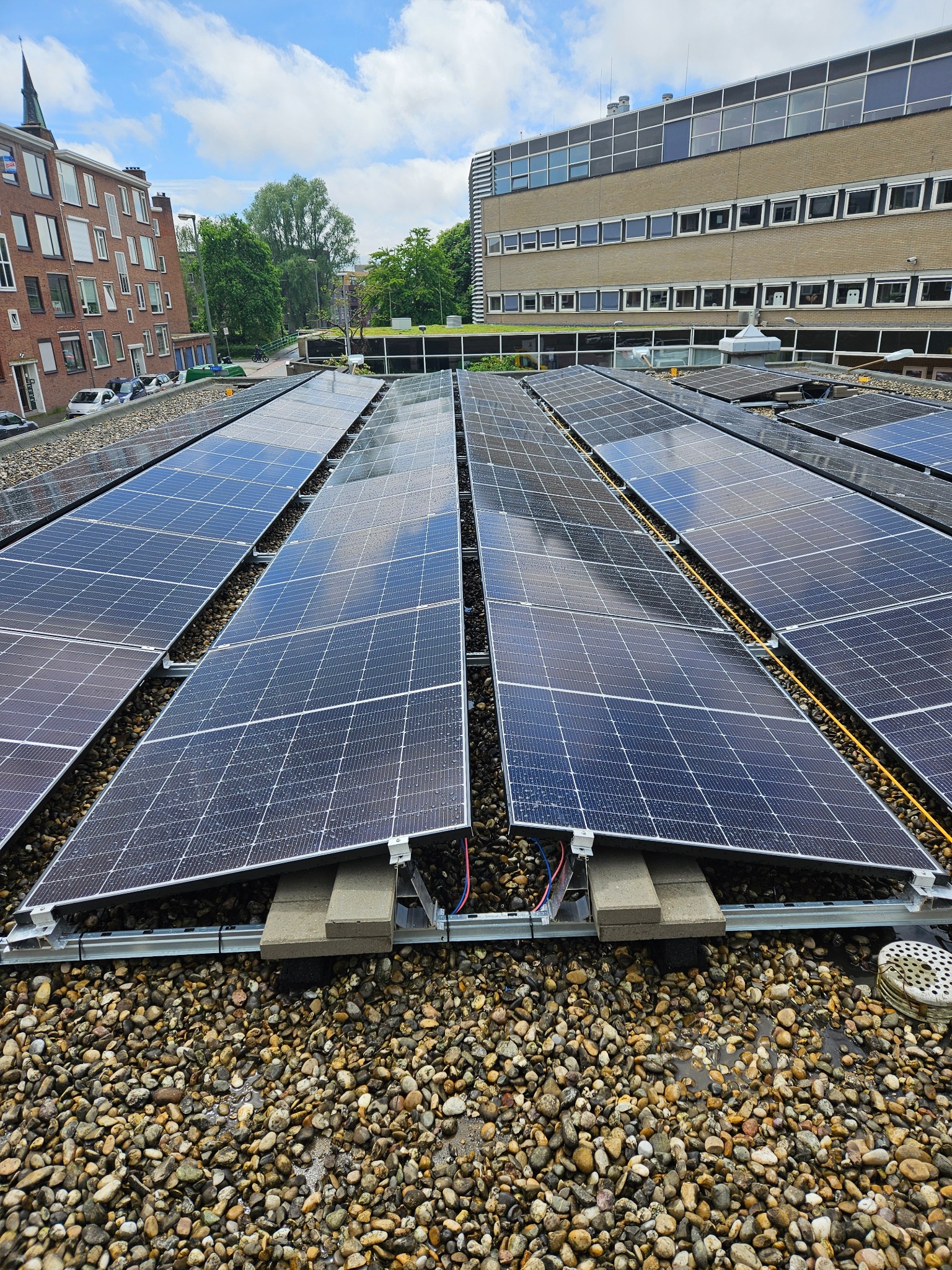 Instalacja fotowoltaiczna na dachu budynku pokrytym drobnymi kamieniami, widoczne panele, kable i system montażowy, w tle budynki mieszkalne i biurowe.