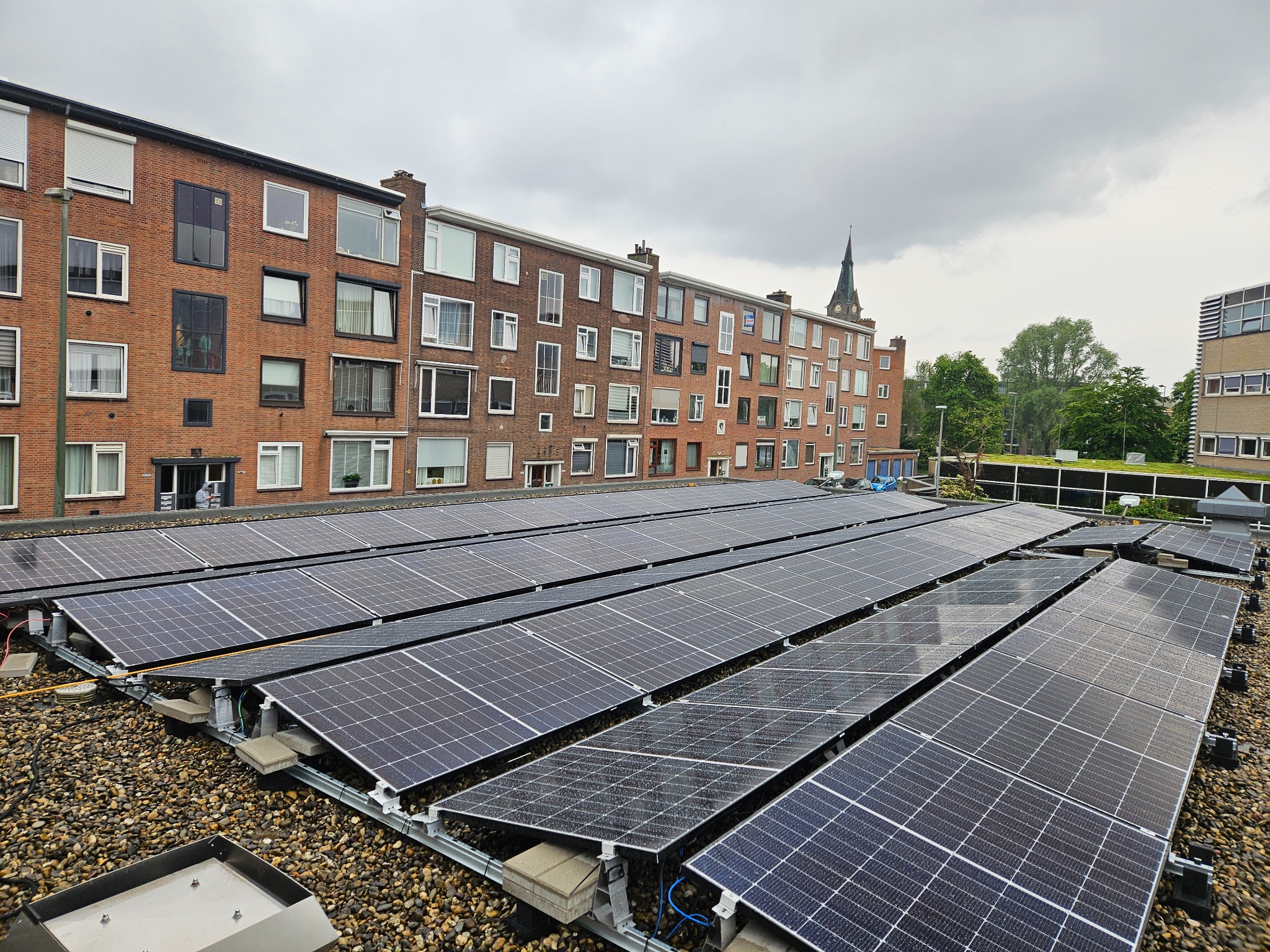 Rząd paneli słonecznych na płaskim dachu budynku mieszkalnego z cegły, widok z góry, w tle wieża kościelna.