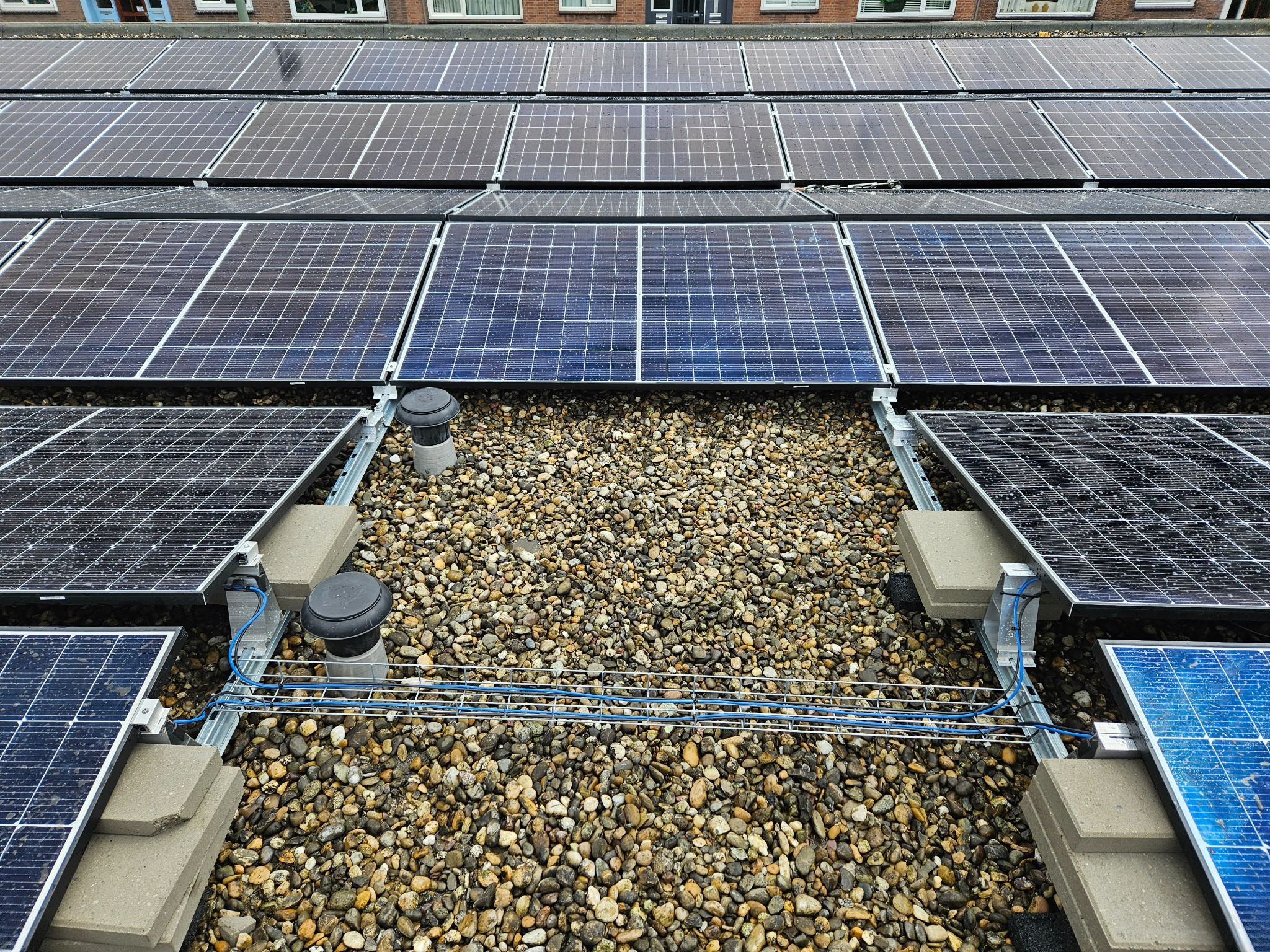 Instalacja paneli słonecznych na płaskim dachu pokrytym drobnymi kamieniami, widoczne elementy montażowe i okablowanie.