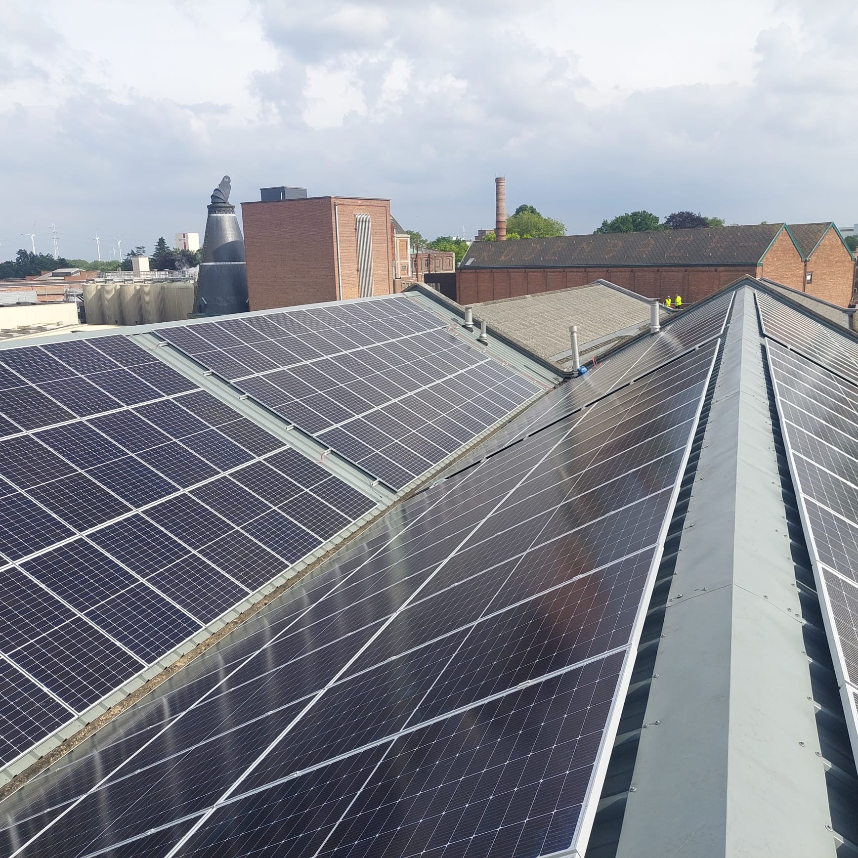 Rozległa instalacja paneli słonecznych na dachu budynku przemysłowego, z widocznymi kominami i innymi budynkami w tle, oraz dwoma osobami w żółtych ubraniach na dachu.