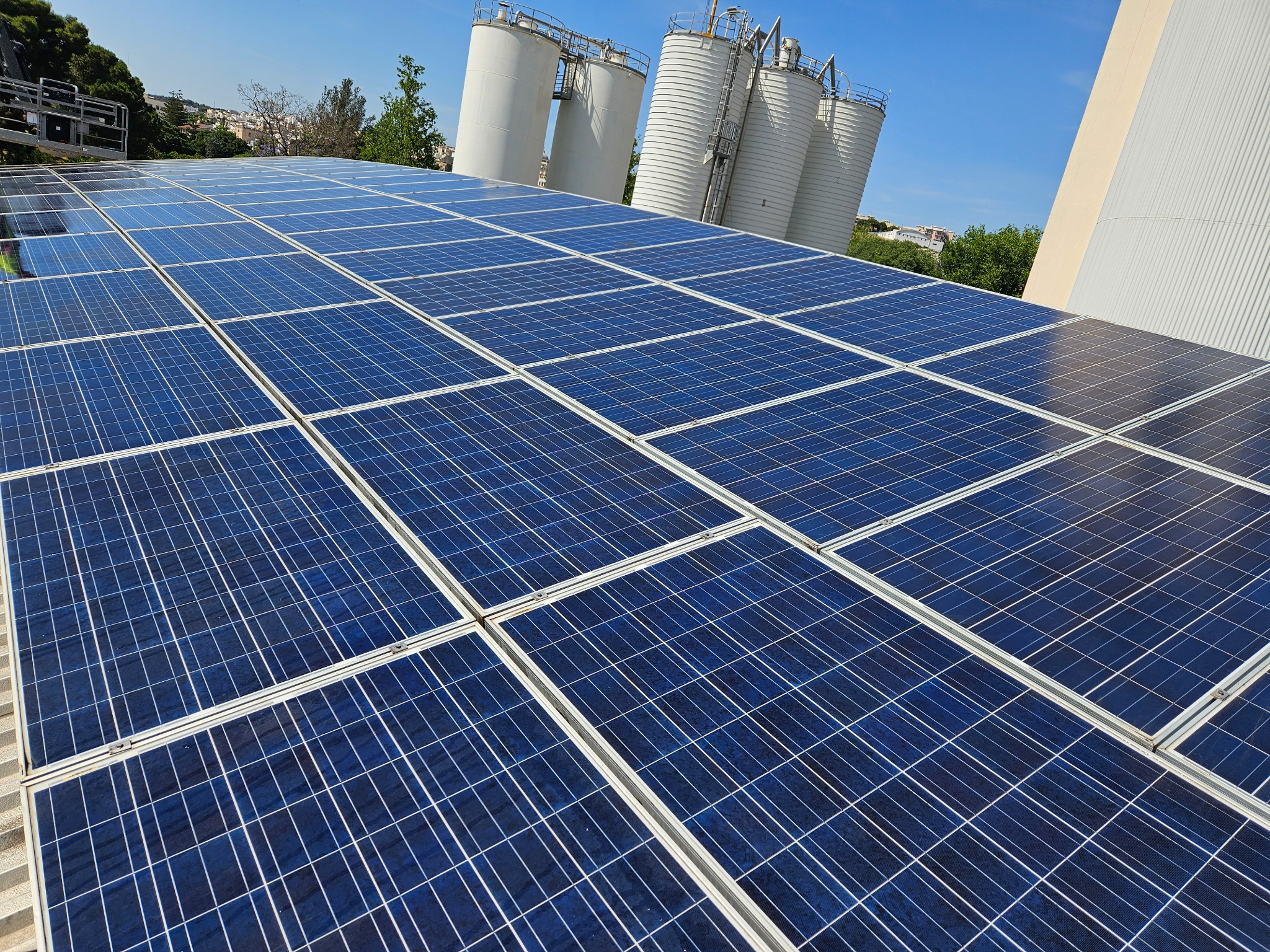 Instalacja fotowoltaiczna na dachu budynku przemysłowego, w tle silosy i błękitne niebo.