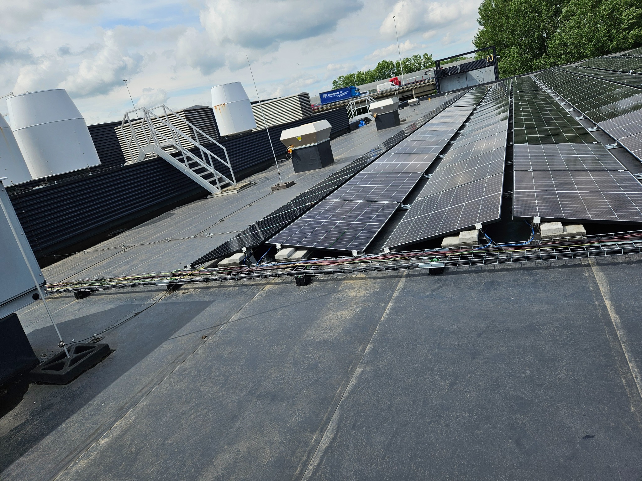 Instalacja paneli słonecznych na dachu przemysłowym z widocznymi elementami wentylacyjnymi i kablami w korytkach.