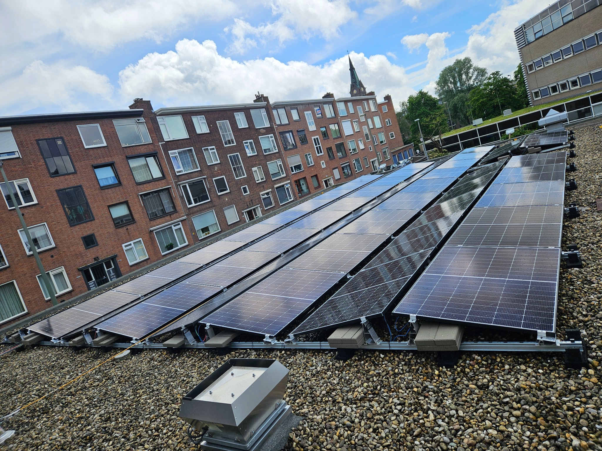 Rząd paneli słonecznych na płaskim dachu pokrytym żwirem, w tle budynek mieszkalny z cegły i wieża kościelna pod błękitnym niebem z chmurami.