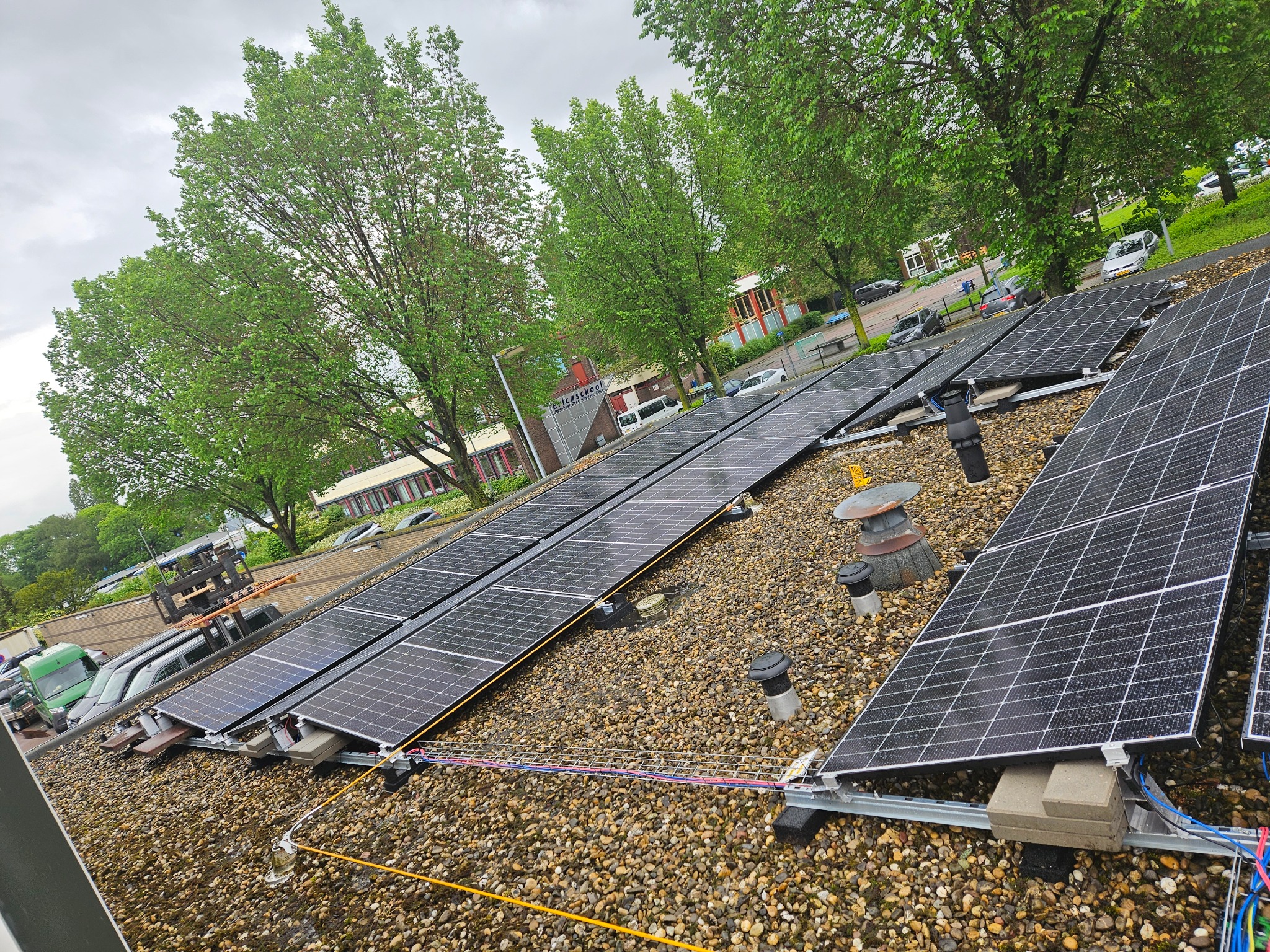 Instalacja paneli słonecznych na dachu pokrytym drobnym żwirem, widoczne kable i elementy montażowe, w tle zieleń drzew i budynek szkoły.