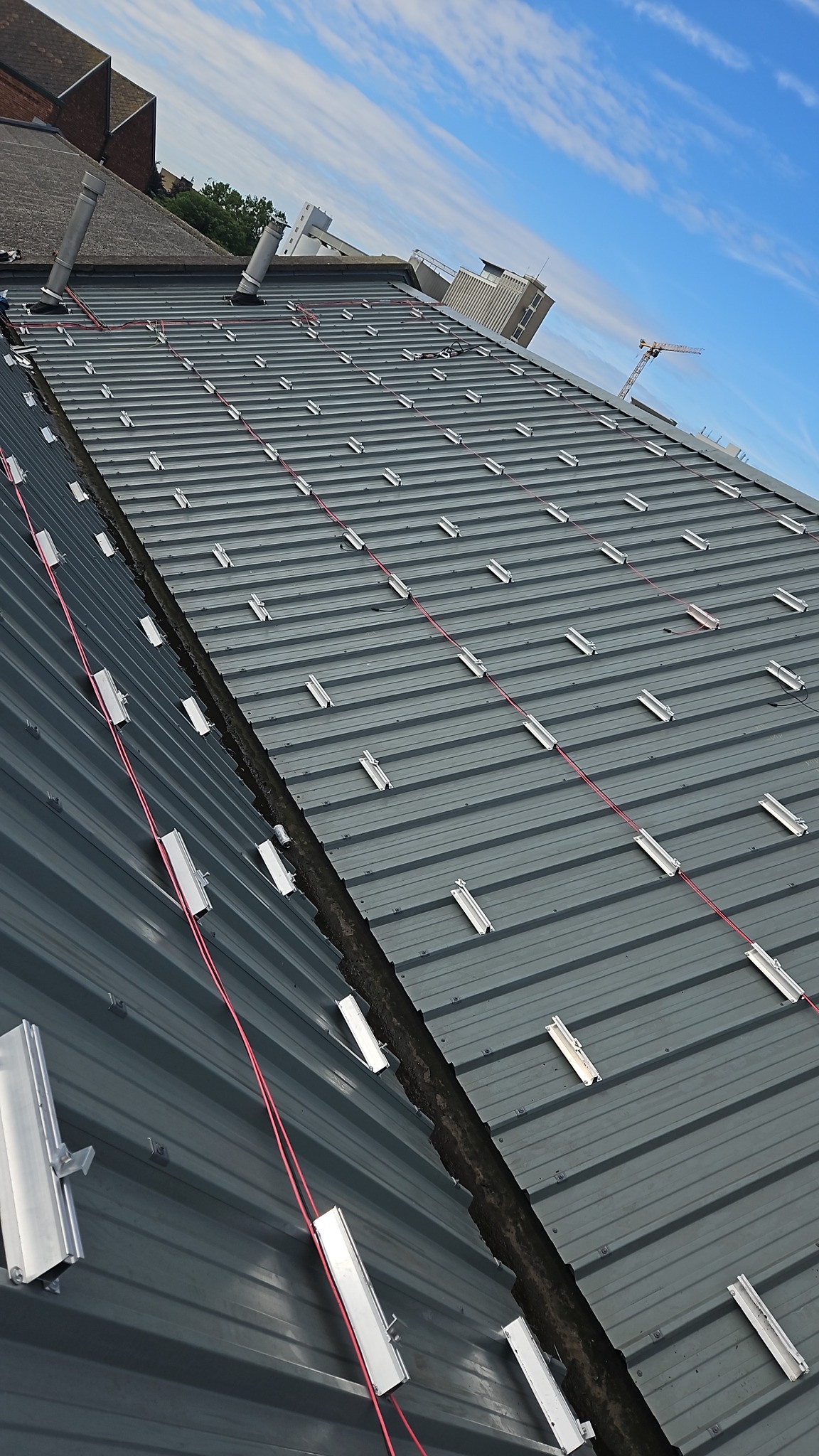 Montaż systemu szynowego pod panele słoneczne na dachu z blachy trapezowej, widoczne kable i elementy mocujące, w tle budynki i błękitne niebo.