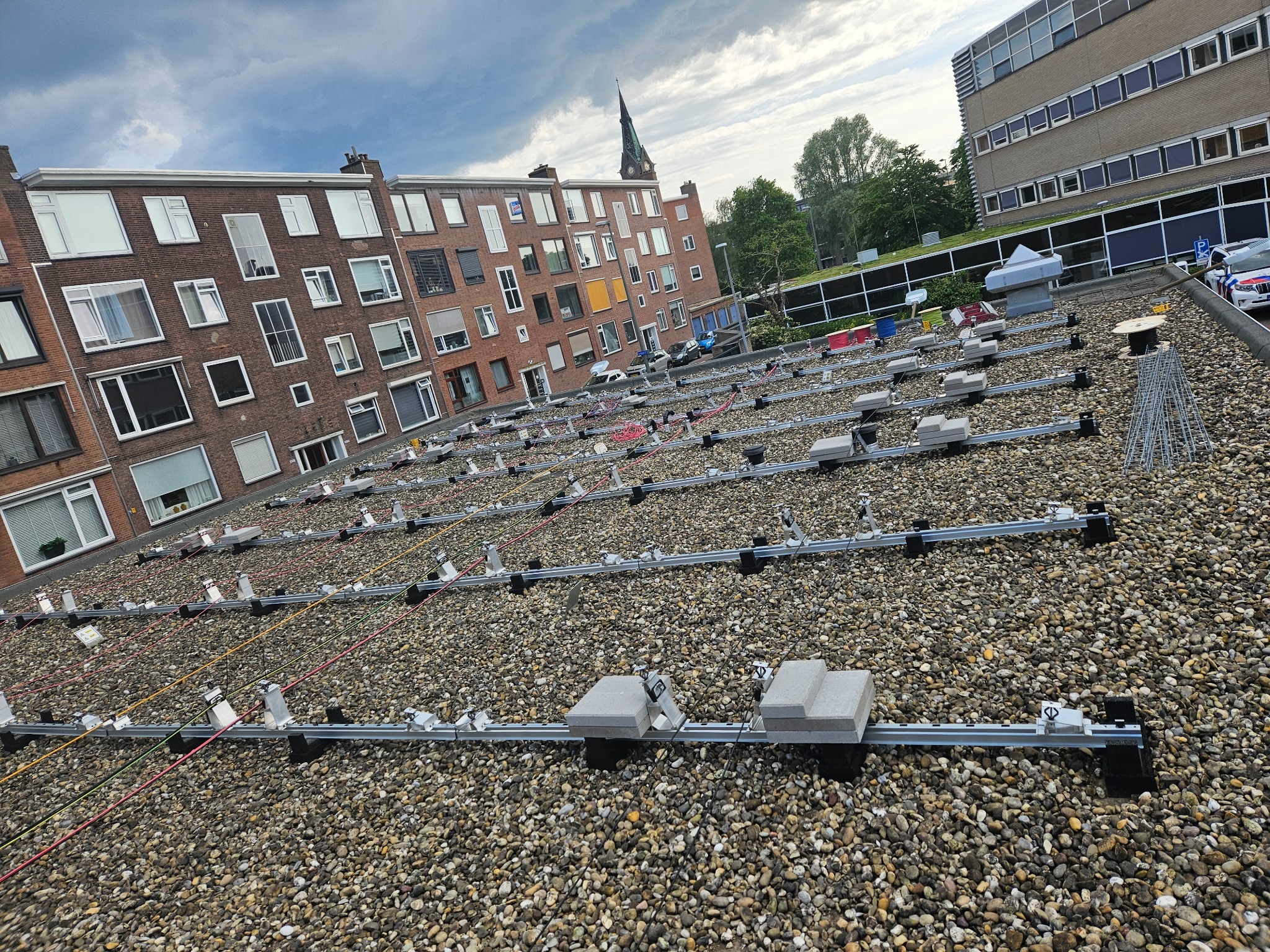 Montaż konstrukcji wsporczej pod panele fotowoltaiczne na dachu pokrytym drobnym żwirem, widoczne przewody, w tle budynki mieszkalne i kościół.