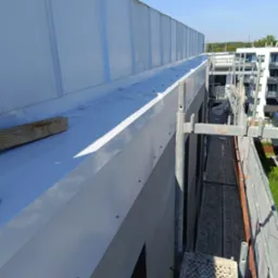 Montaż obróbki blacharskiej na dachu budynku wielorodzinnego, widoczne rusztowanie i fragmenty elewacji w tle.