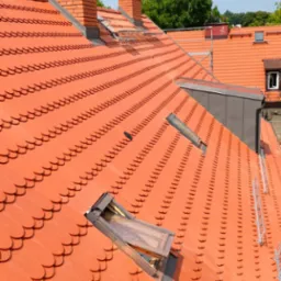 Czerwona dachówka karpiówka na dachu z widocznymi kominami z cegły, otwartym oknem dachowym i fragmentem rusztowania w tle, w słoneczny dzień.