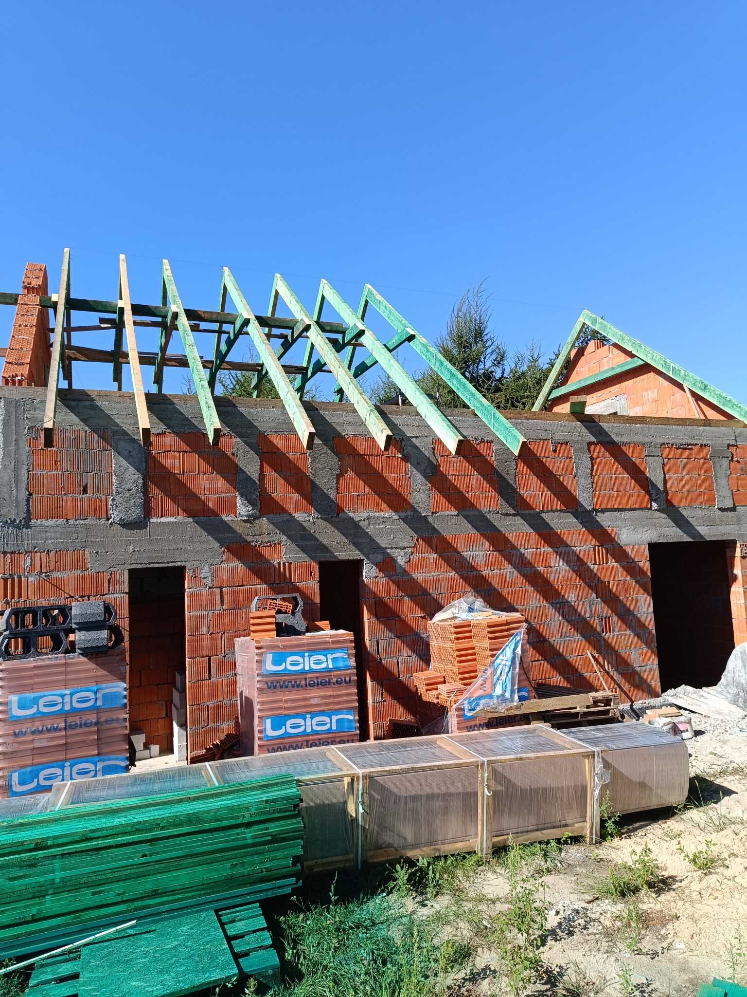 Budowa domu jednorodzinnego w stanie surowym otwartym: ściany z czerwonej cegły, zielona konstrukcja więźby dachowej, palety z materiałami budowlanymi Leier na pierwszym planie, błękitne niebo w tle.