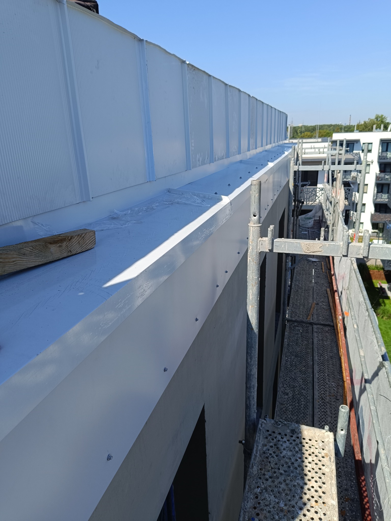 Montaż obróbki blacharskiej na dachu budynku wielorodzinnego, widoczne rusztowanie i fragmenty elewacji w tle.