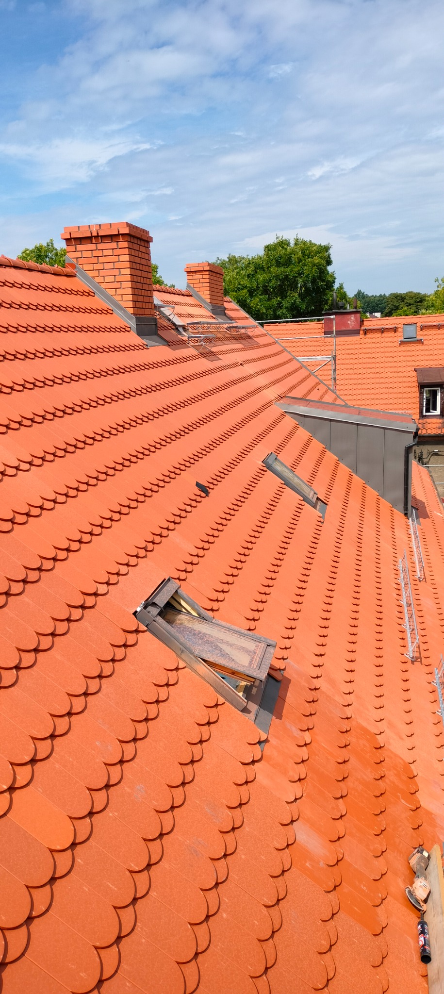 Czerwona dachówka karpiówka na dachu z widocznymi kominami z cegły, otwartym oknem dachowym i fragmentem rusztowania w tle, w słoneczny dzień.