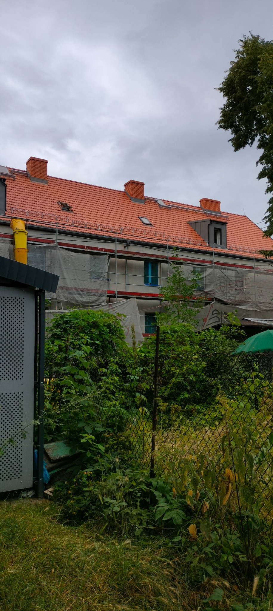 Dwupiętrowy dom w trakcie prac remontowych, widoczne rusztowania osłonięte siatką, nowa dachówka w kolorze ceglastym, częściowo zasłonięty bujną roślinnością od strony ogrodu.