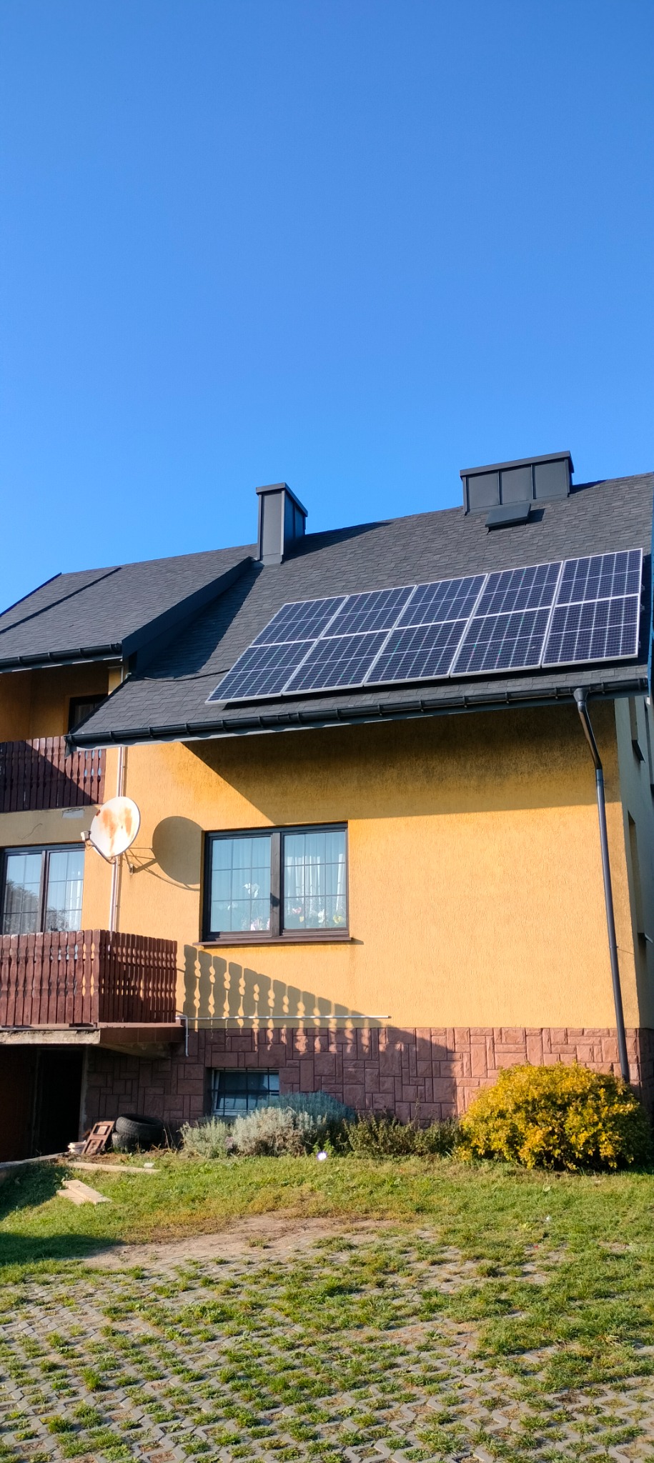 Żółty dom jednorodzinny z szarym dachem i panelami słonecznymi, widoczne elementy elewacji i balkonu, antena satelitarna na ścianie.