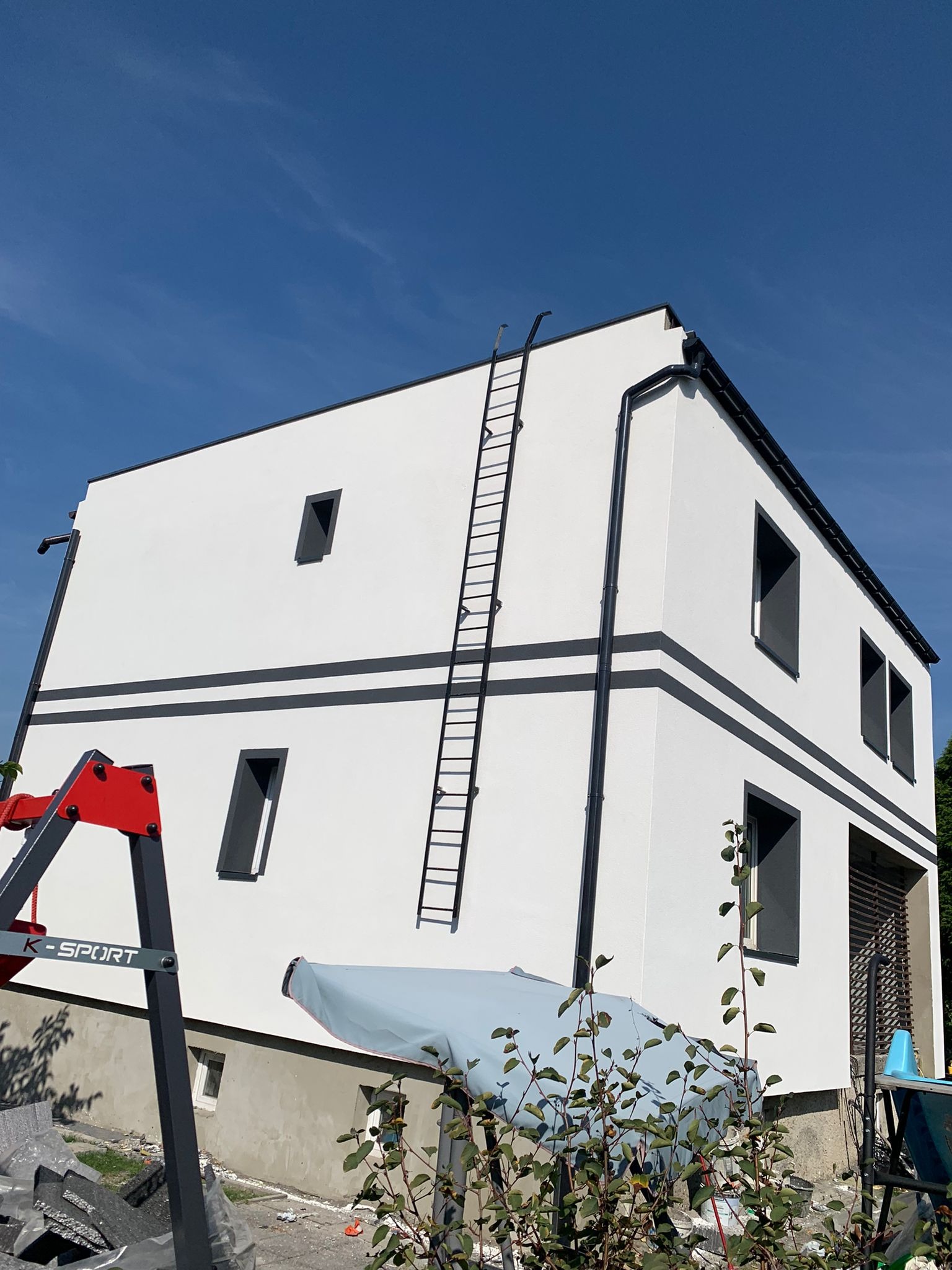 Nowoczesny dom z białą elewacją, szarymi detalami i oknami, z drabiną opartą o ścianę, widok z dołu na tle błękitnego nieba.