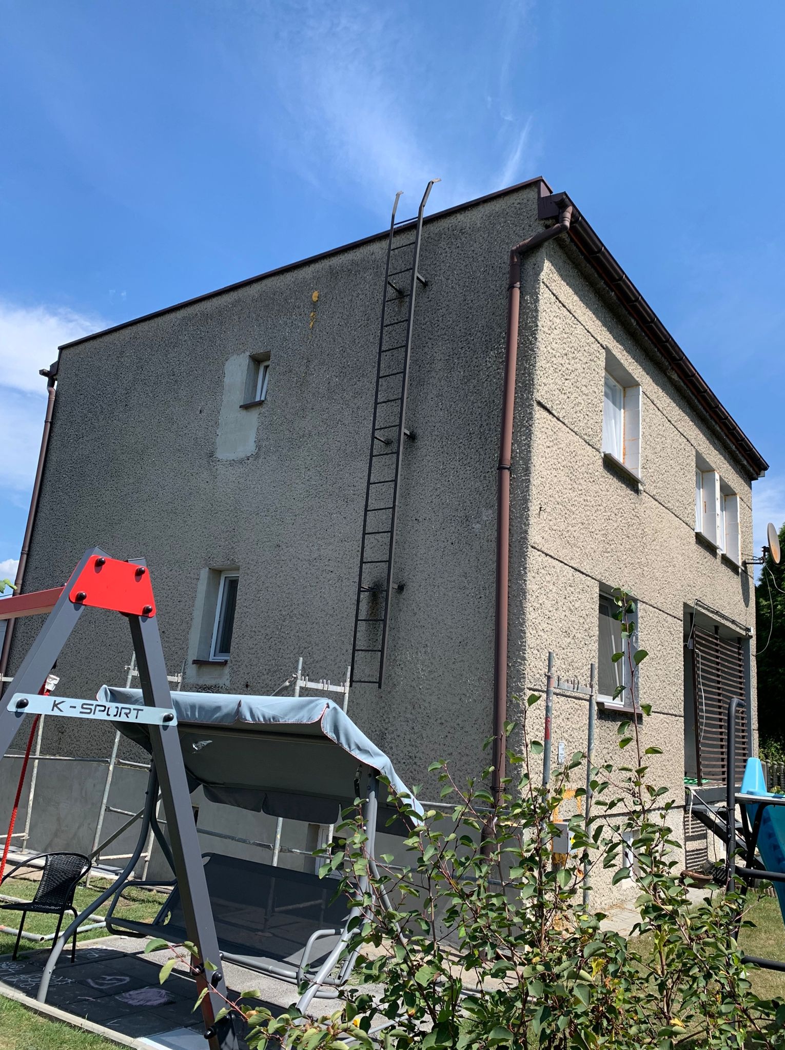 Dwupiętrowy dom z szarą elewacją o fakturze baranka, z metalową drabiną przymocowaną do ściany, widoczne ślady napraw elewacji wokół okna, huśtawka ogrodowa na pierwszym planie.