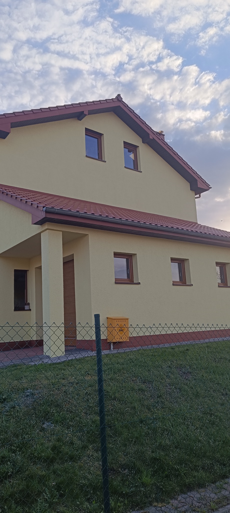 Jasny dom jednorodzinny z brązowym dachem i stolarką okienną, widziany zza zielonego ogrodzenia. Żółta skrzynka gazowa przy elewacji.