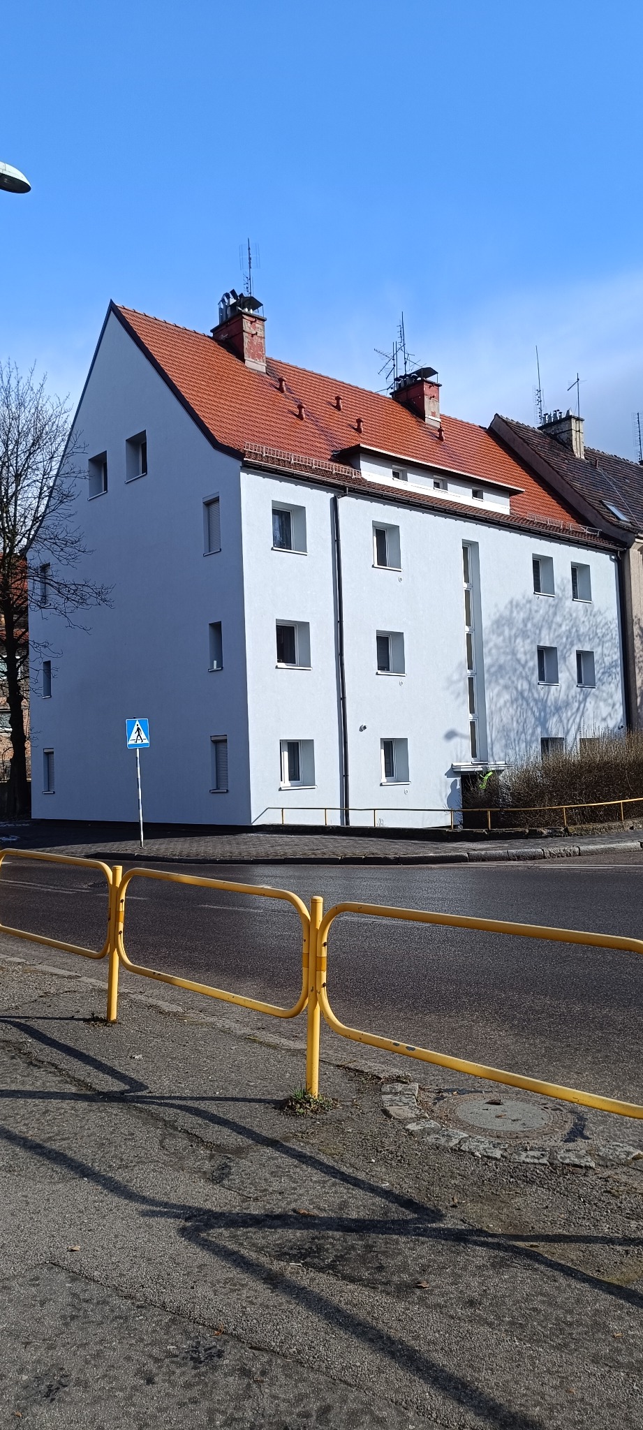 Odnowiony budynek wielorodzinny z czerwoną dachówką, widoczny z poziomu ulicy, z żółtą barierką i znakiem drogowym. Jasne, słoneczne oświetlenie.