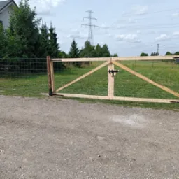 Brama z surowego drewna z zamkiem, wkomponowana w ogrodzenie z siatki na tle zielonej łąki i słupa wysokiego napięcia.