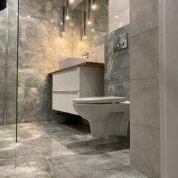 Mateusz Zakrzewski - Nowoczesna łazienka z szarymi płytkami imitującymi beton, wisząca toaleta, biała szafka z umywalką na drewnianym blacie i minimalistyczne lampy wiszące.