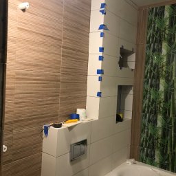 Mateusz Zakrzewski - Białe płytki z niebieską taśmą maskującą podczas montażu w łazience, widoczne narzędzia i fragment wanny, na ścianie płytki imitujące drewno i dekoracyjne z motywem bambusa.