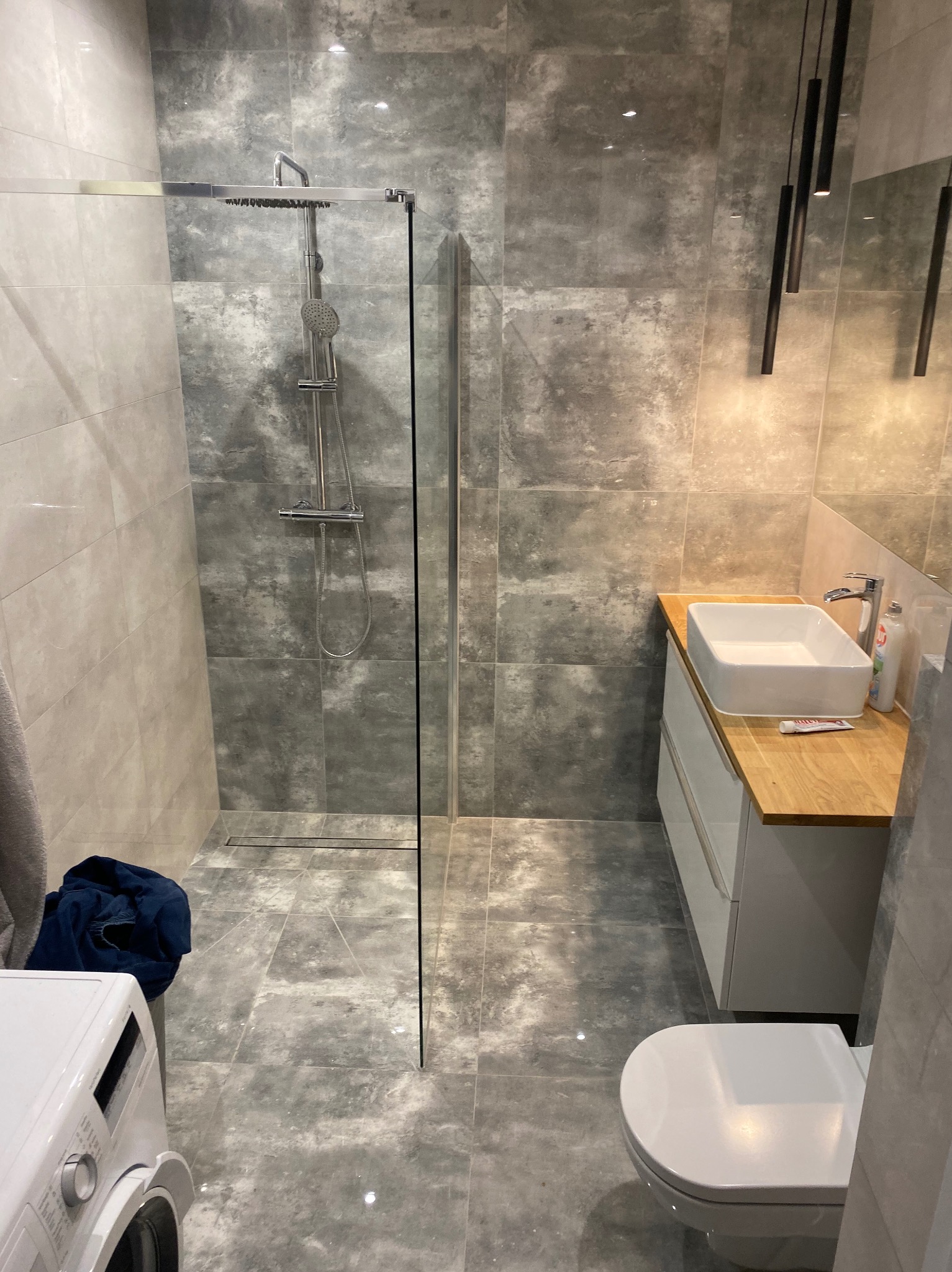 Nowoczesna łazienka we Wrocławiu z szarymi płytkami imitującymi beton, kabiną prysznicową typu walk-in, umywalką nablatową na drewnianym blacie i wiszącą toaletą.