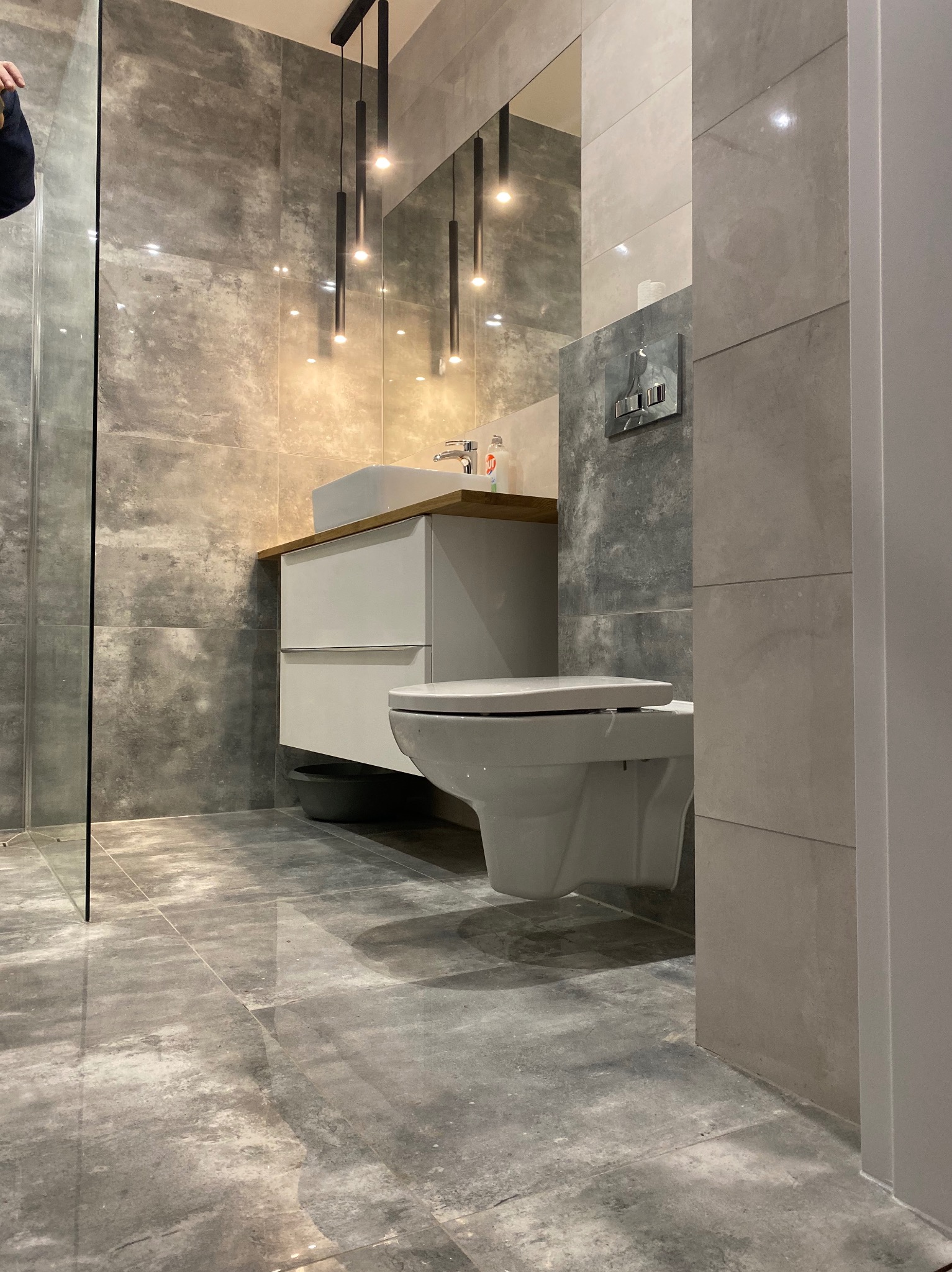 Nowoczesna łazienka z szarymi płytkami imitującymi beton, wisząca toaleta, biała szafka z umywalką na drewnianym blacie i minimalistyczne lampy wiszące.