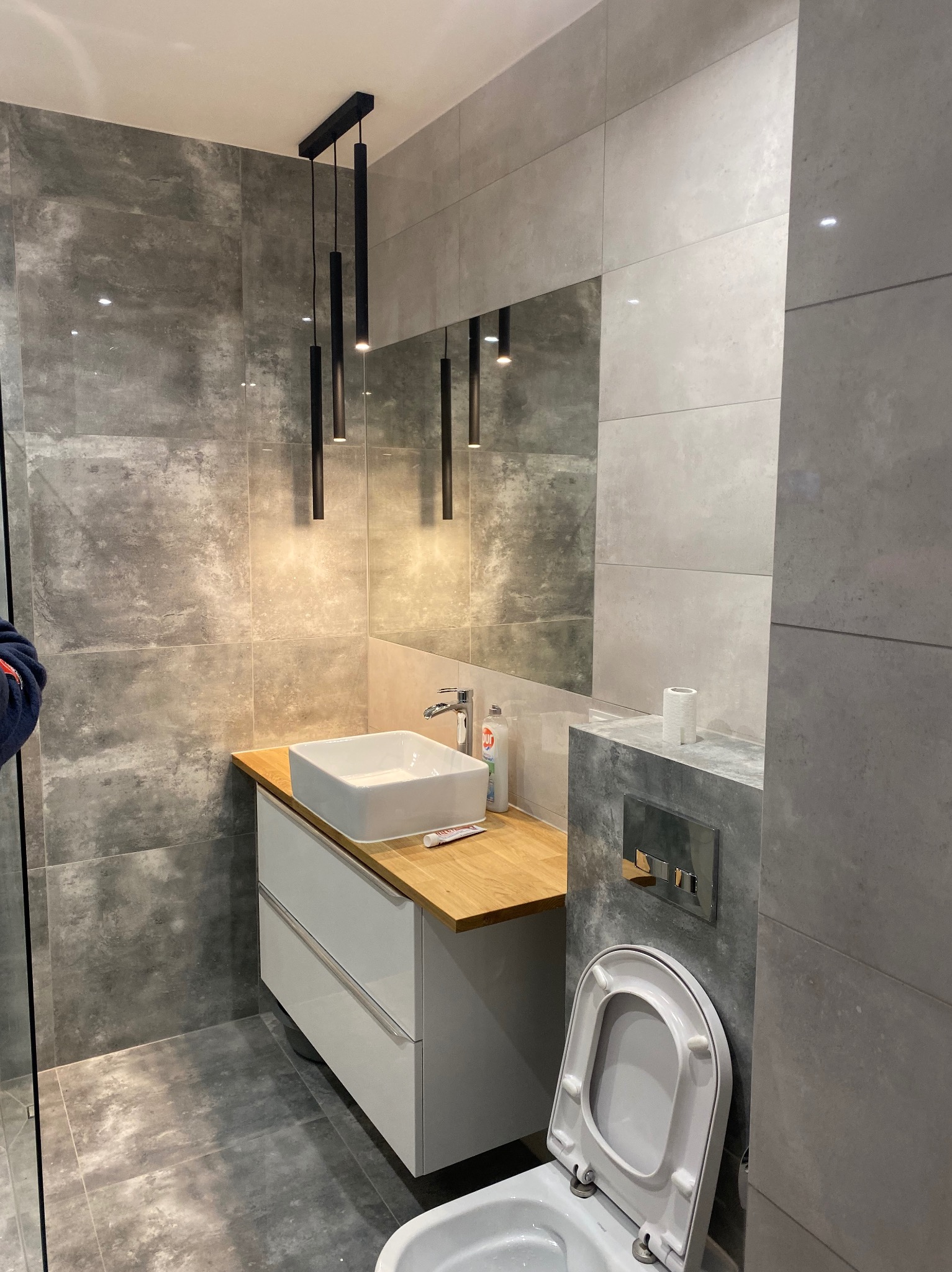 Nowoczesna łazienka z szarymi płytkami, wiszącą szafką z umywalką na drewnianym blacie i minimalistycznym oświetleniem.