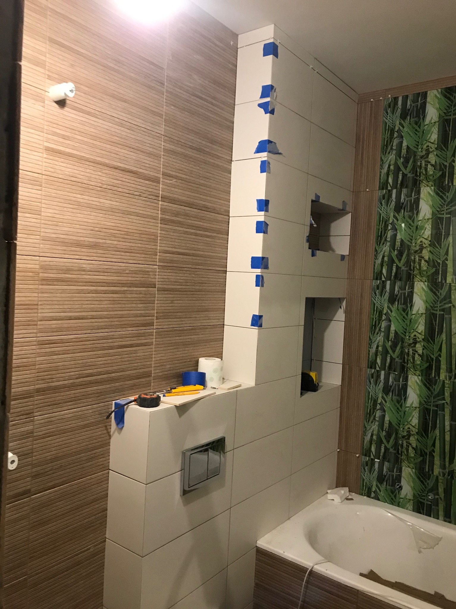 Białe płytki z niebieską taśmą maskującą podczas montażu w łazience, widoczne narzędzia i fragment wanny, na ścianie płytki imitujące drewno i dekoracyjne z motywem bambusa.