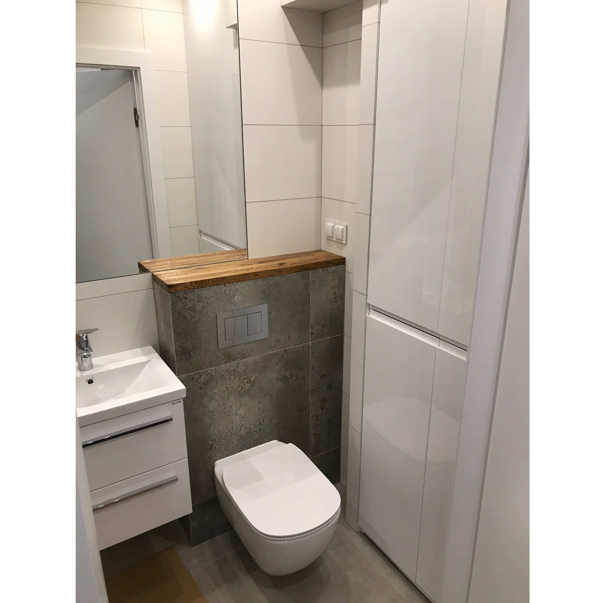Nowoczesna łazienka z białymi płytkami, umywalką z szafką Elita, podwieszaną toaletą, zabudową meblową i drewnianym blatem pod lustrem.