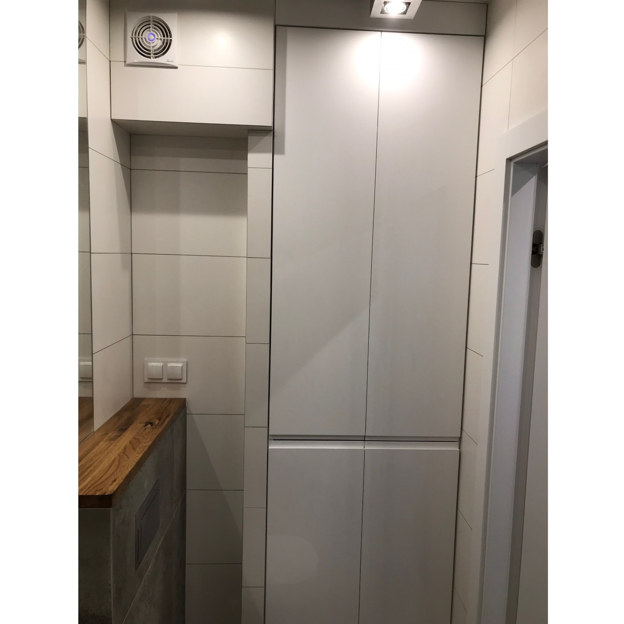 Nowoczesna łazienka z białymi płytkami, drewnianym blatem umywalki i zabudową meblową pod sufit, z widocznym wentylatorem i oświetleniem punktowym.