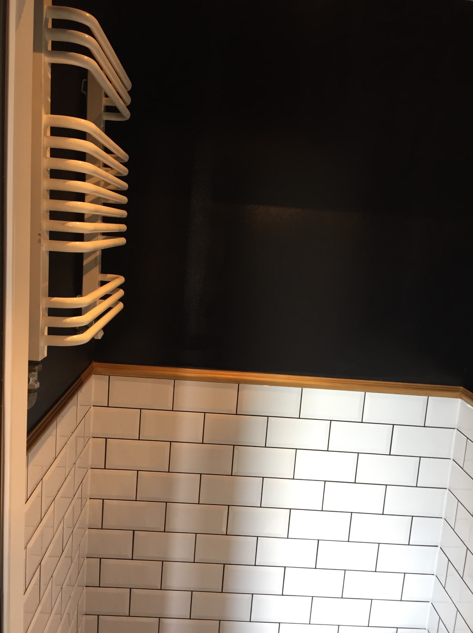 Nowoczesna łazienka z czarną ścianą i białymi, prostokątnymi płytkami ułożonymi w cegiełkę, wykończona drewnianą listwą i wyposażona w biały, drabinkowy grzejnik.