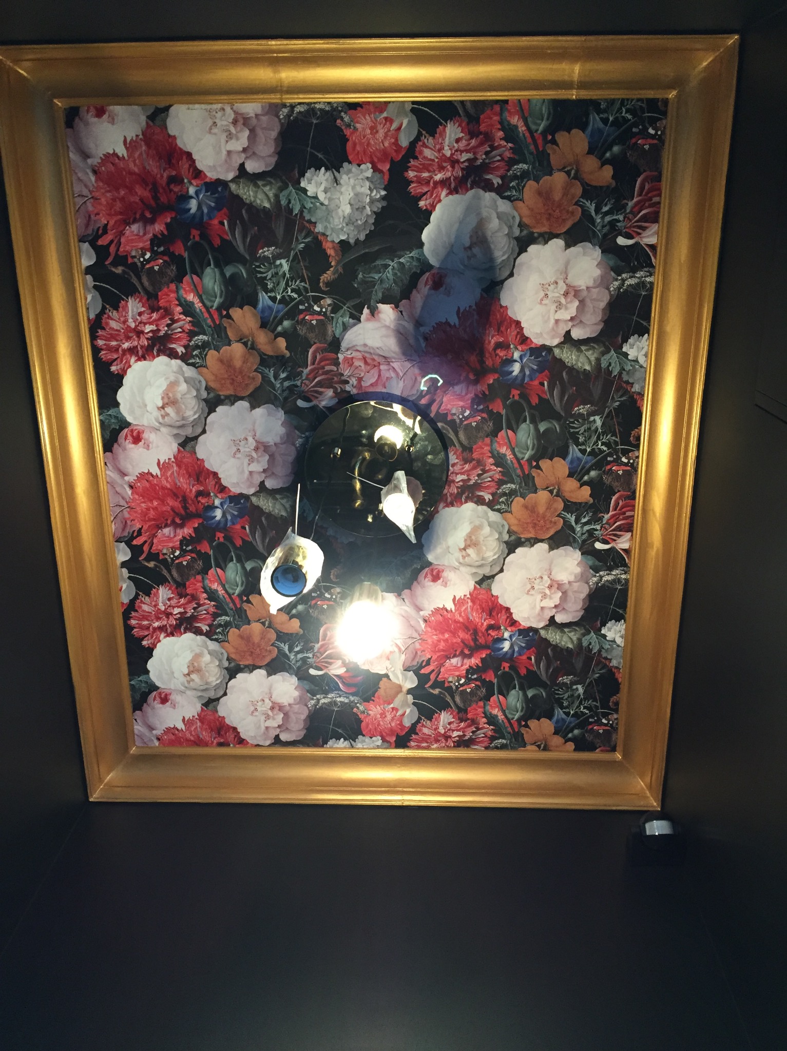 Dekoracyjny panel sufitowy z motywem kwiatowym w złotej ramie, oświetlony lampą wiszącą, na tle ciemnej ściany.