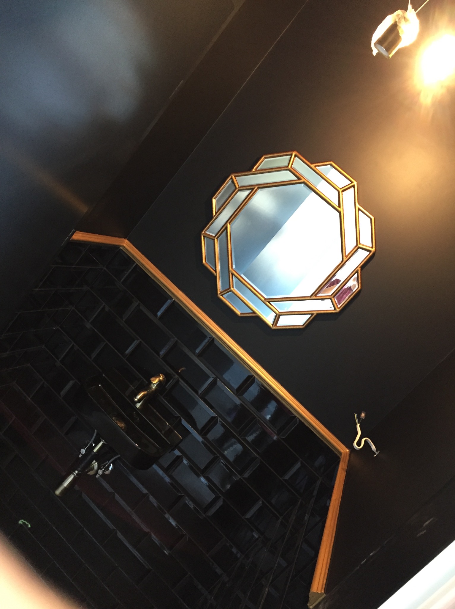 Ciemna łazienka z czarnymi, błyszczącymi płytkami ułożonymi w jodełkę, złotym obramowaniem, umywalką z baterią w kolorze złota i ozdobnym lustrem w geometrycznej ramie.