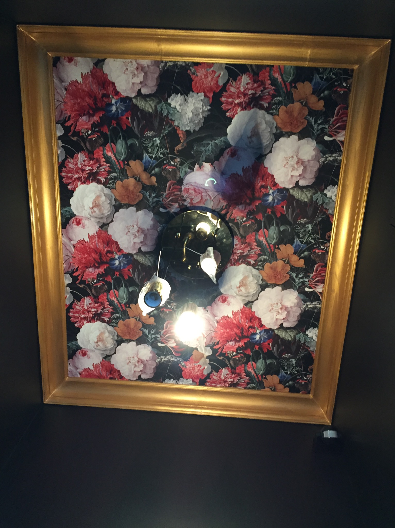 Kaseton z kwiecistą tapetą w złotej ramie, oświetlony lampą wiszącą z niebieskim akcentem, zamontowany na ciemnej ścianie.