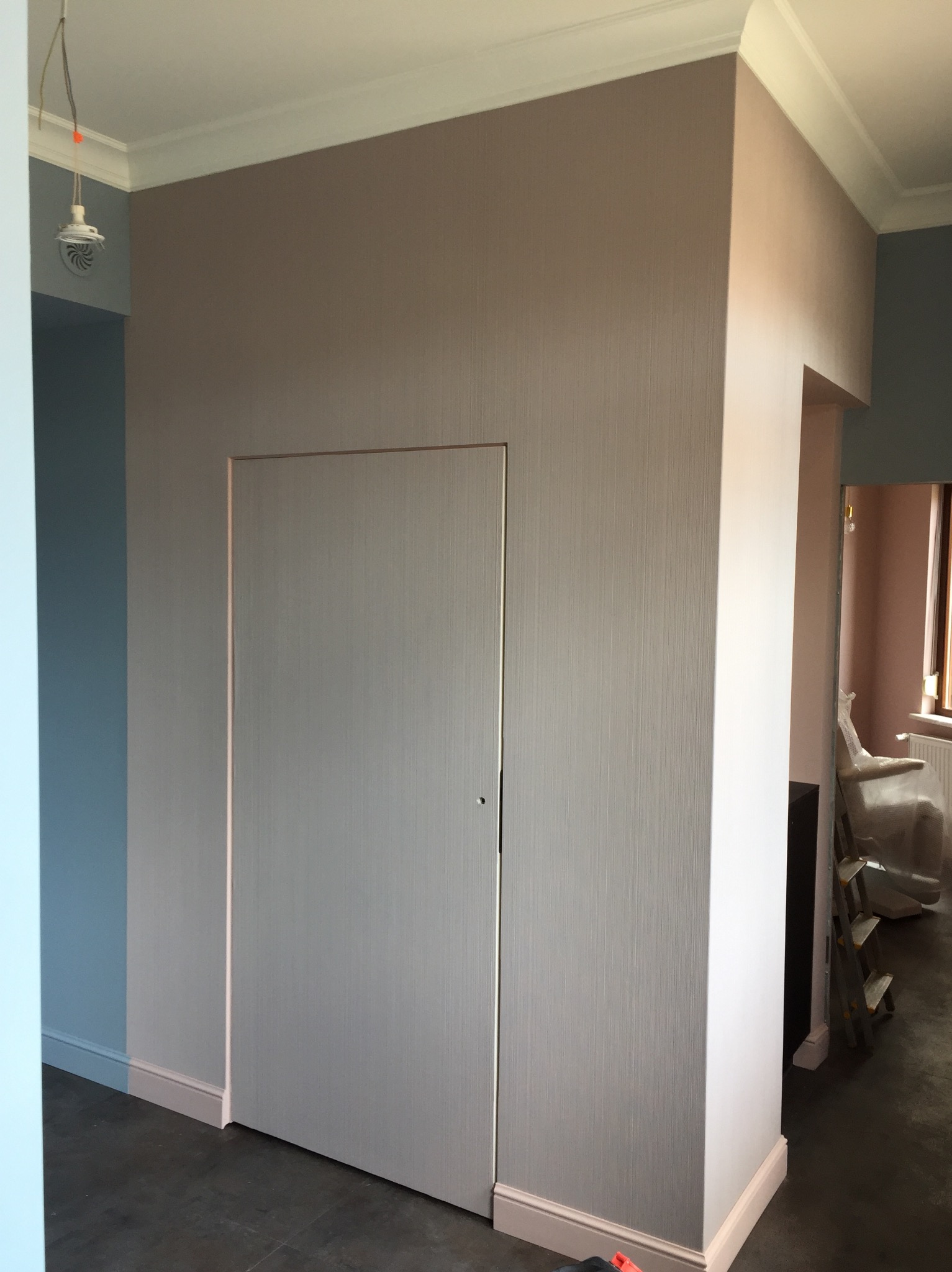 Pudrowo-różowe ściany po remoncie z ukrytymi drzwiami, widoczna listwa przypodłogowa i sufitowa, oraz fragment instalacji oświetleniowej.