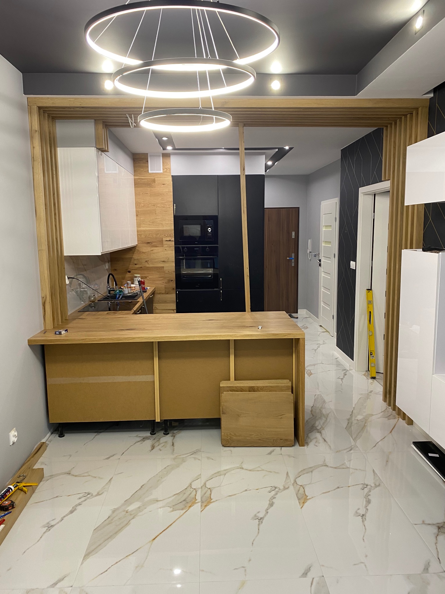 Wnętrze mieszkania w trakcie prac wykończeniowych: widoczna drewniana konstrukcja oddzielająca kuchnię od salonu, białe szafki kuchenne, marmurowe płytki podłogowe i poziomica oparta o ścianę.