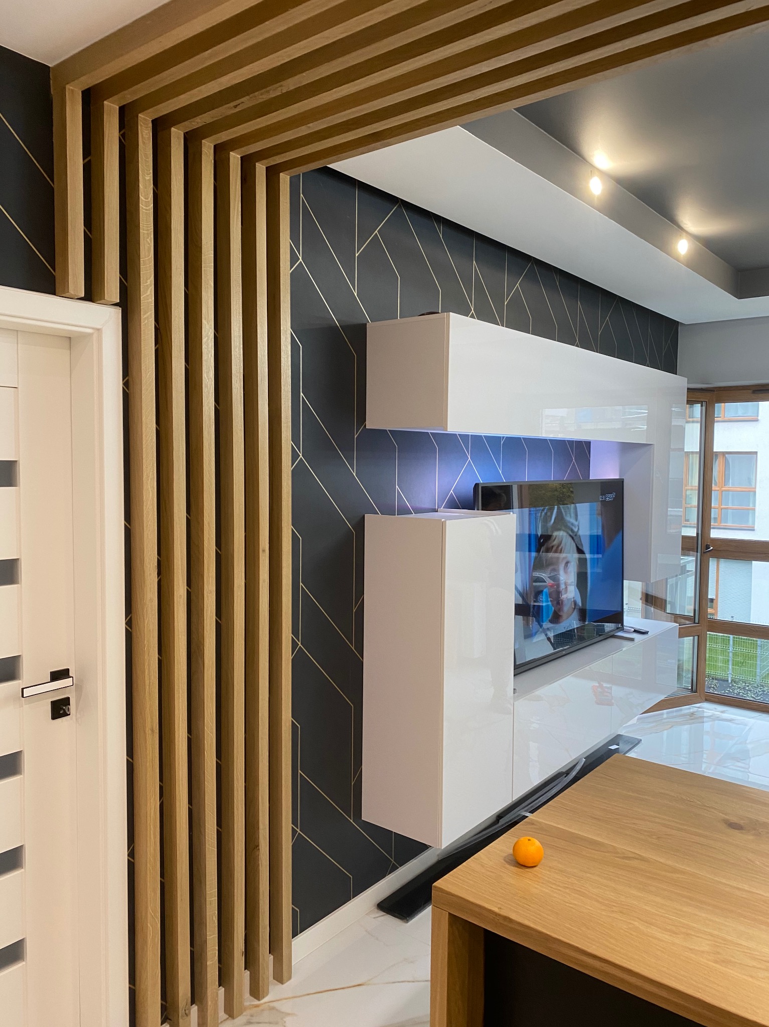 Nowoczesne wnętrze: drewniana ażurowa ścianka działowa przy białych drzwiach, ściana w geometryczne wzory, białe meble na wysoki połysk z podświetleniem LED, marmurowa podłoga i drewniany stół...