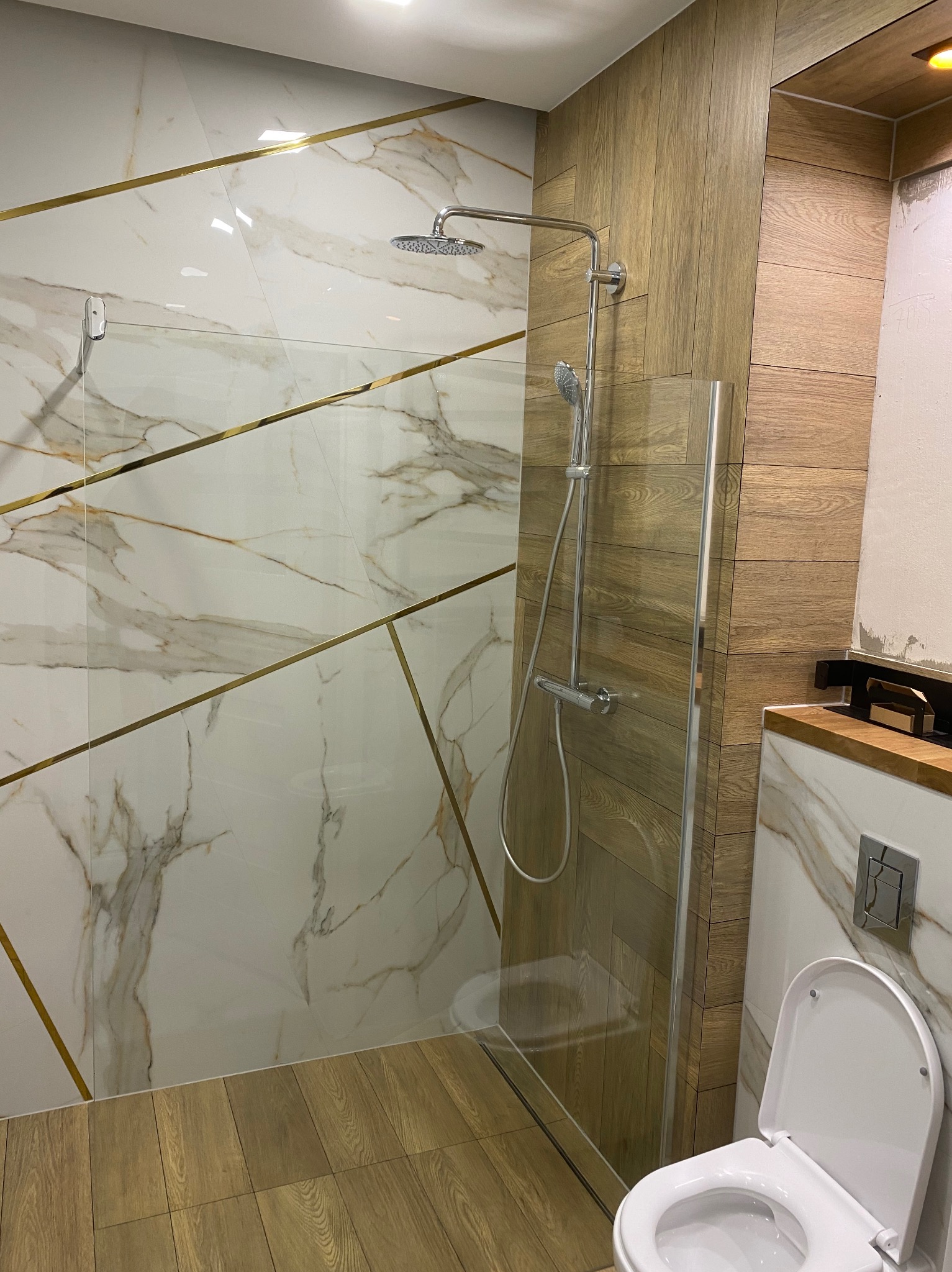 Nowoczesna kabina prysznicowa z deszczownicą i szklaną ścianką, wykończona białymi płytkami imitującymi marmur z geometrycznym wzorem złotych pasków, oraz drewnopodobnymi panelami ściennymi...