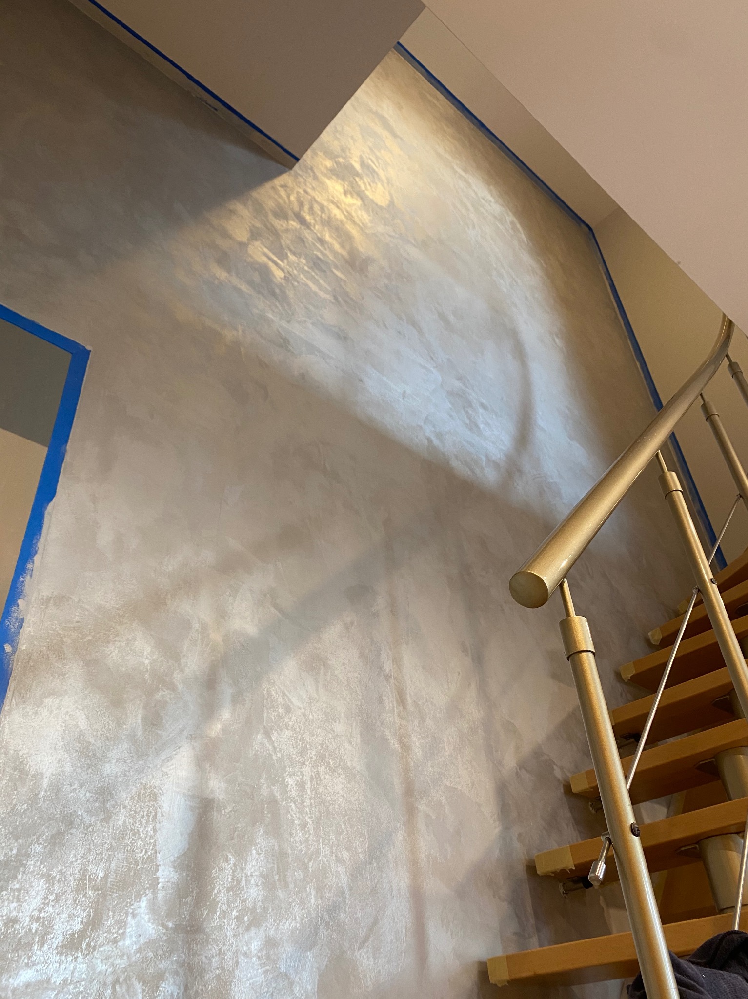 Ściana pokryta dekoracyjną, srebrzystą farbą z efektem perłowym, zabezpieczona niebieską taśmą malarską przy suficie i framudze drzwi, widoczne schody z metalową balustradą.