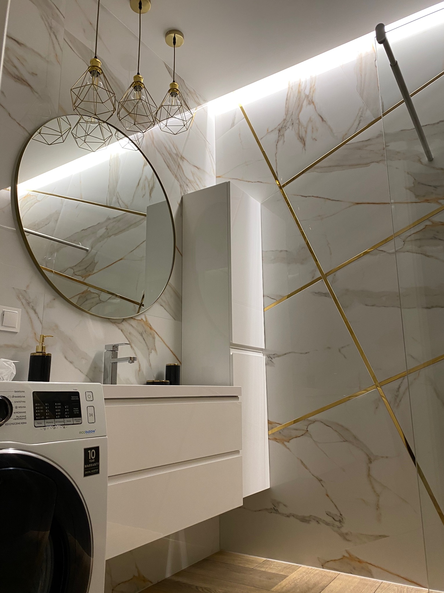Nowoczesna łazienka z marmurowymi płytkami zdobionymi złotymi listwami, okrągłym lustrem z oświetleniem LED, białymi szafkami pod umywalką i pralką Samsung z 10-letnią gwarancją na silnik Digital...