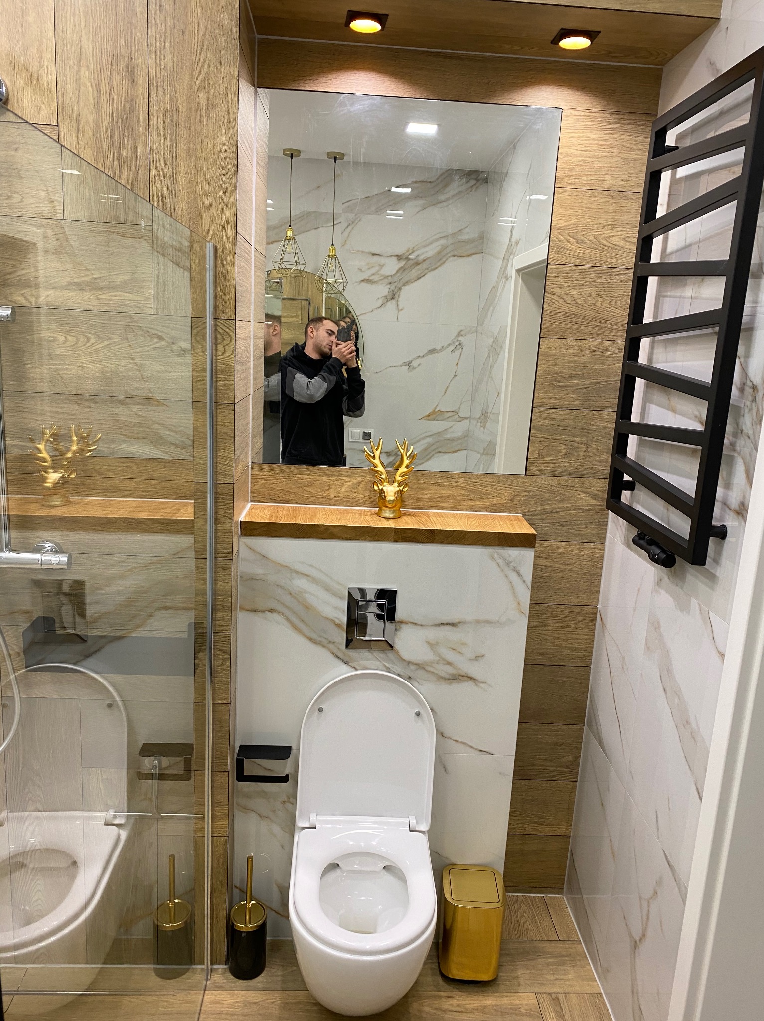 Nowoczesna łazienka z elementami drewna i marmuru, widoczna kabina prysznicowa ze złotymi akcentami oraz toaleta z połyskującym złotym koszem na śmieci; odbicie w lustrze osoby robiącej zdjęcie.
