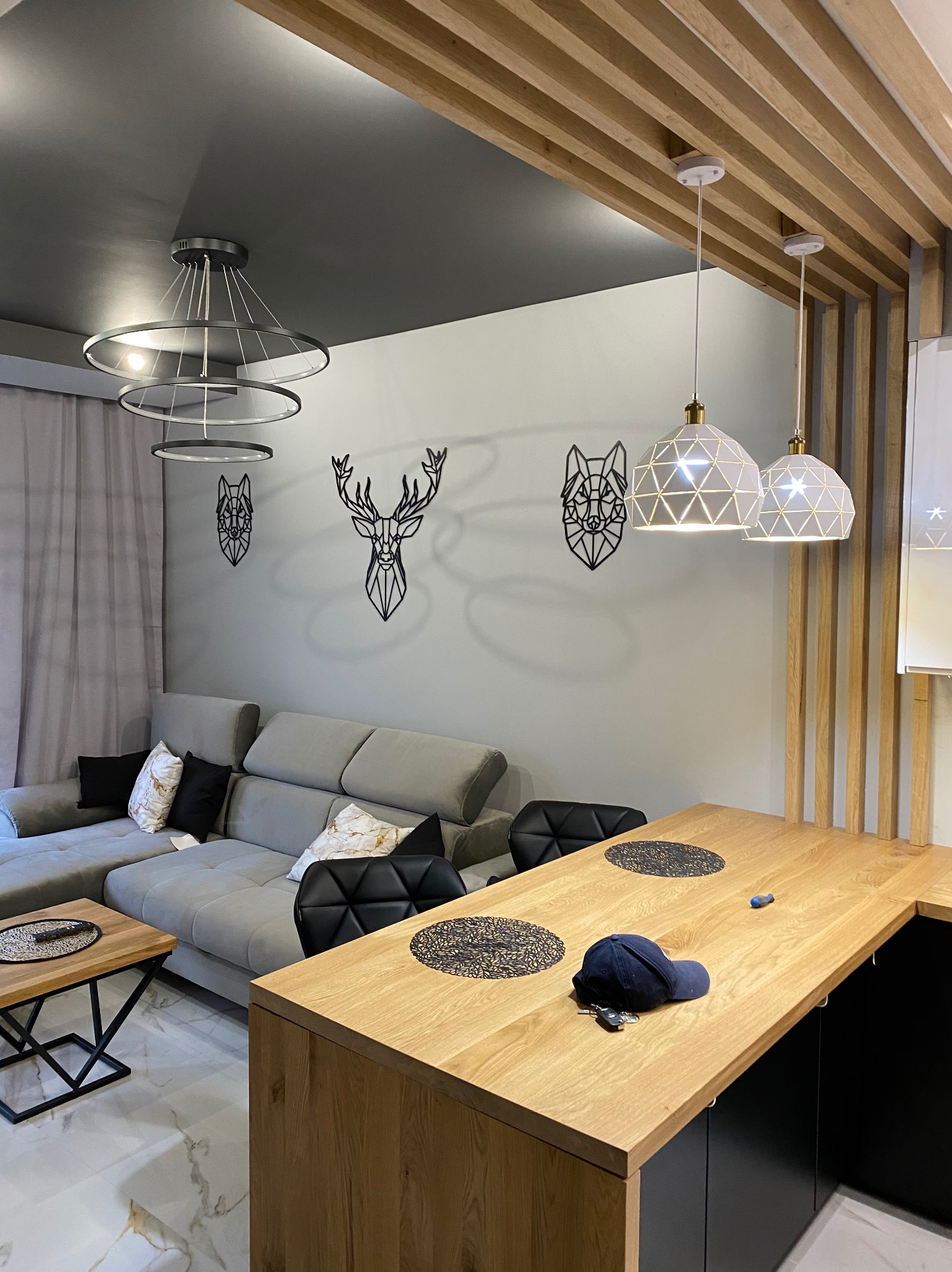 Nowoczesne wnętrze: szara sofa narożna z wzorzystymi poduszkami, drewniany stół z czarnymi akcentami, dekoracje ścienne w kształcie zwierząt i geometryczne lampy wiszące.