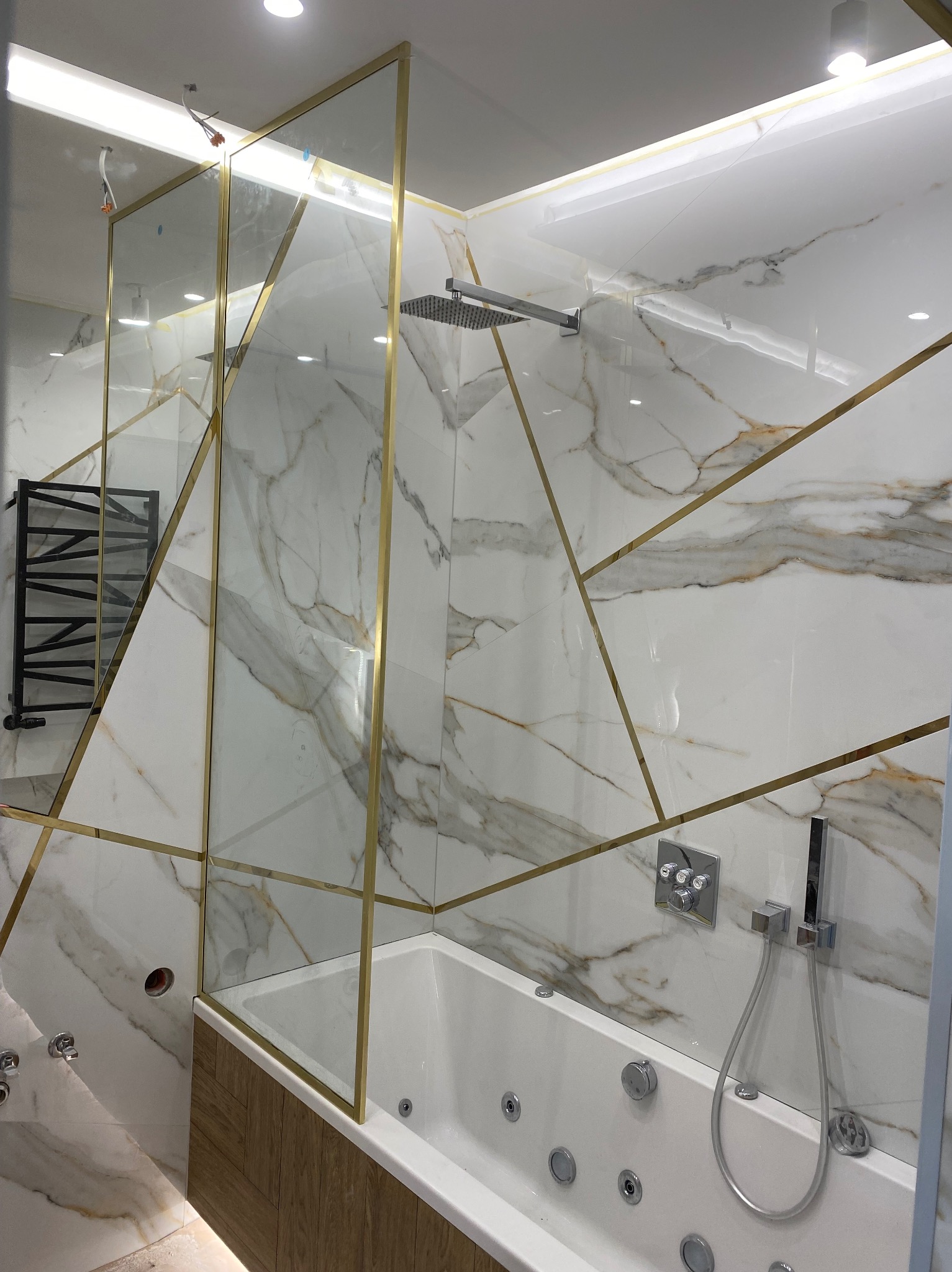 Nowoczesna łazienka z wanną jacuzzi, marmurowymi płytkami z geometrycznymi złotymi wstawkami i szklanym panelem prysznicowym z złotymi obramowaniami.