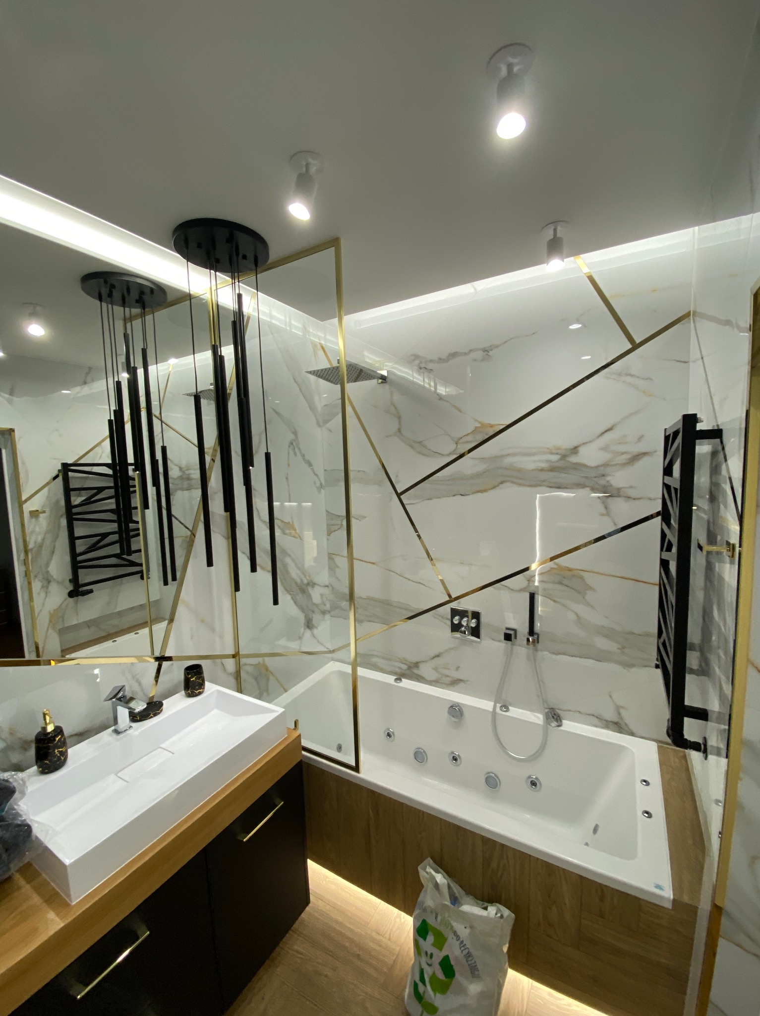 Nowoczesna łazienka z marmurowymi płytkami z geometrycznym złotym wzorem, białą wanną z hydromasażem, umywalką nablatową i czarnymi elementami oświetleniowymi oraz grzejnikiem drabinkowym.