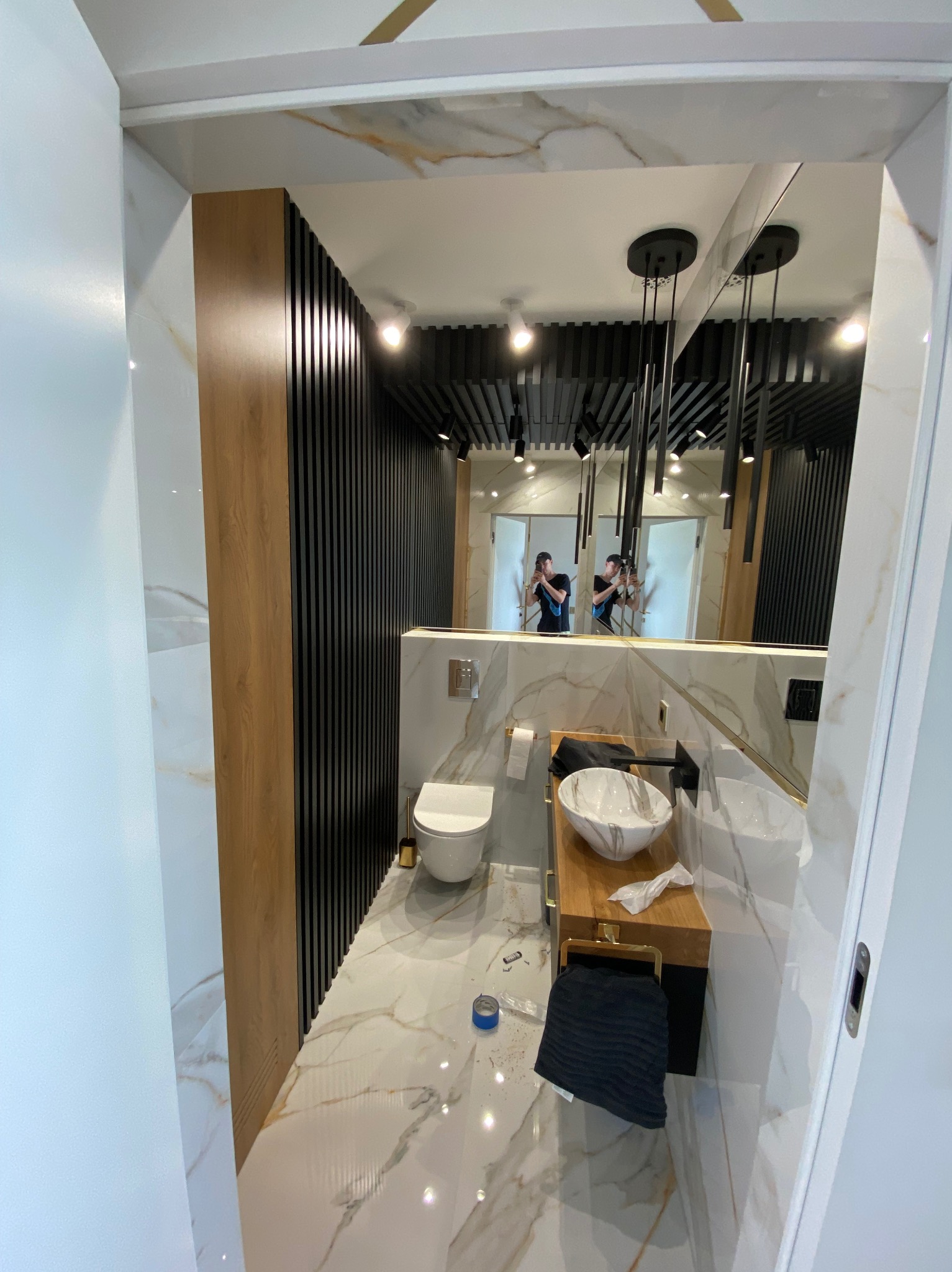 Nowoczesna łazienka z marmurową podłogą i ścianami, umywalką nablatową i czarnymi elementami dekoracyjnymi, widoczne odbicie osoby robiącej zdjęcie w lustrze.