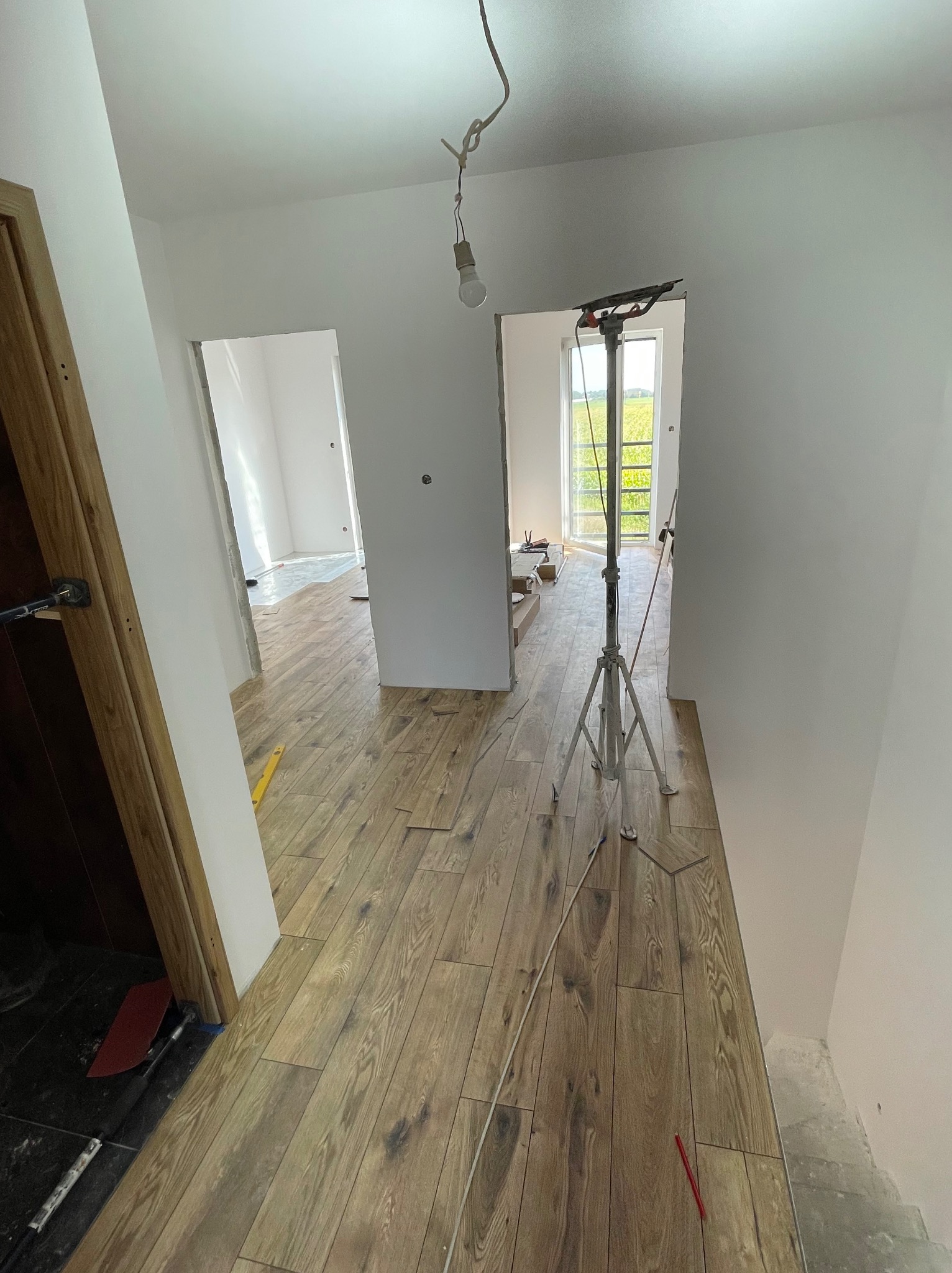 Korytarz w trakcie prac: częściowo ułożone panele podłogowe w odcieniach brązu, widoczne narzędzia, niedokończone ściany i surowa żarówka na kablu.