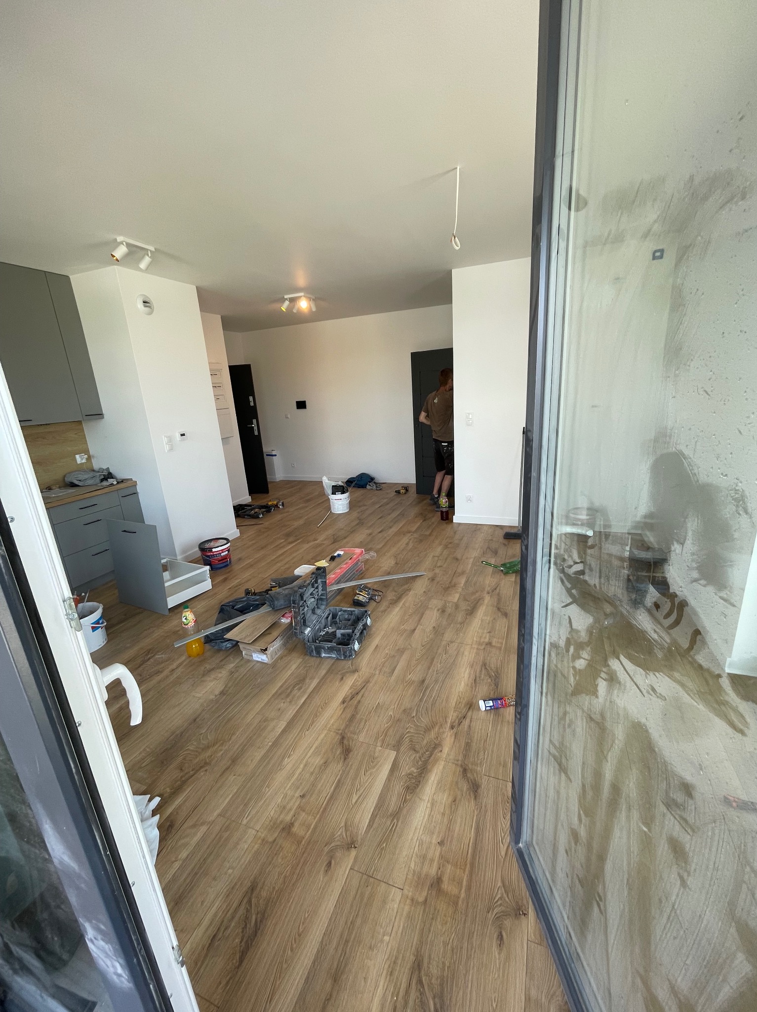 Wnętrze mieszkania w trakcie montażu paneli podłogowych; widoczne narzędzia, częściowo ułożona podłoga, okno balkonowe z zabrudzeniami oraz osoba w tle.